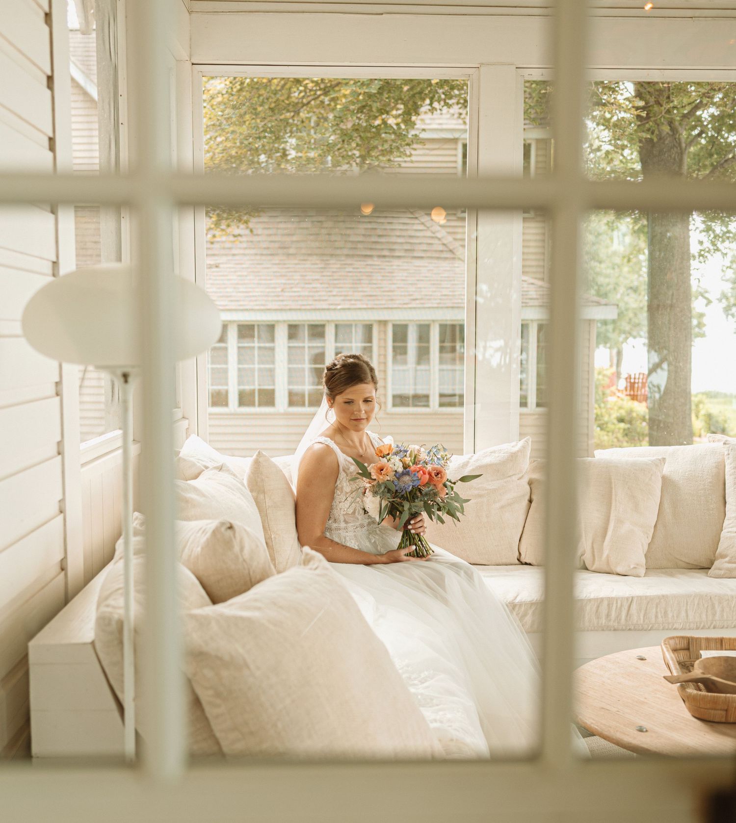 Woman relaxing in bright sunlit room on plush white porch seating with outdoor greenery visible through windows.