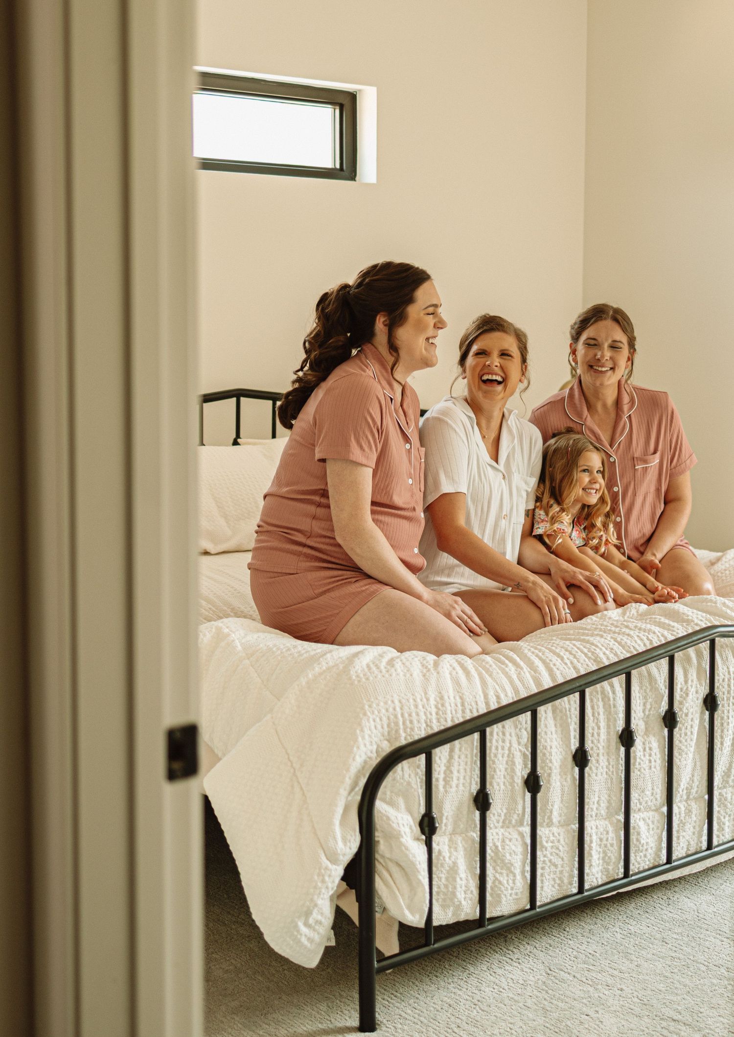 Candid moment of laughter shared on a bed among people in matching pink dresses in a sunlit room.