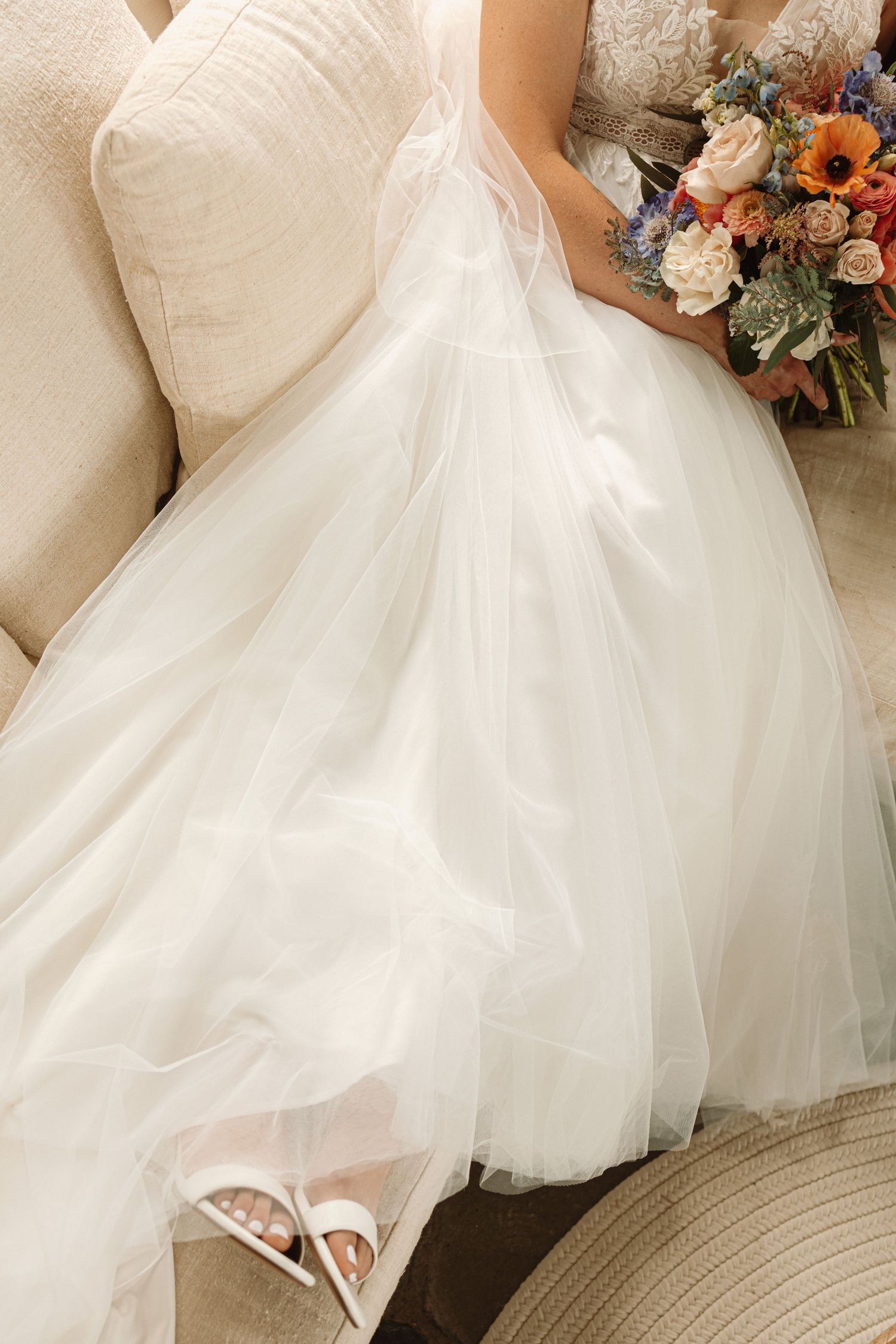 Flowing white tulle wedding dress skirt detail with delicate beaded embellishments and a peach and blue floral bouquet.