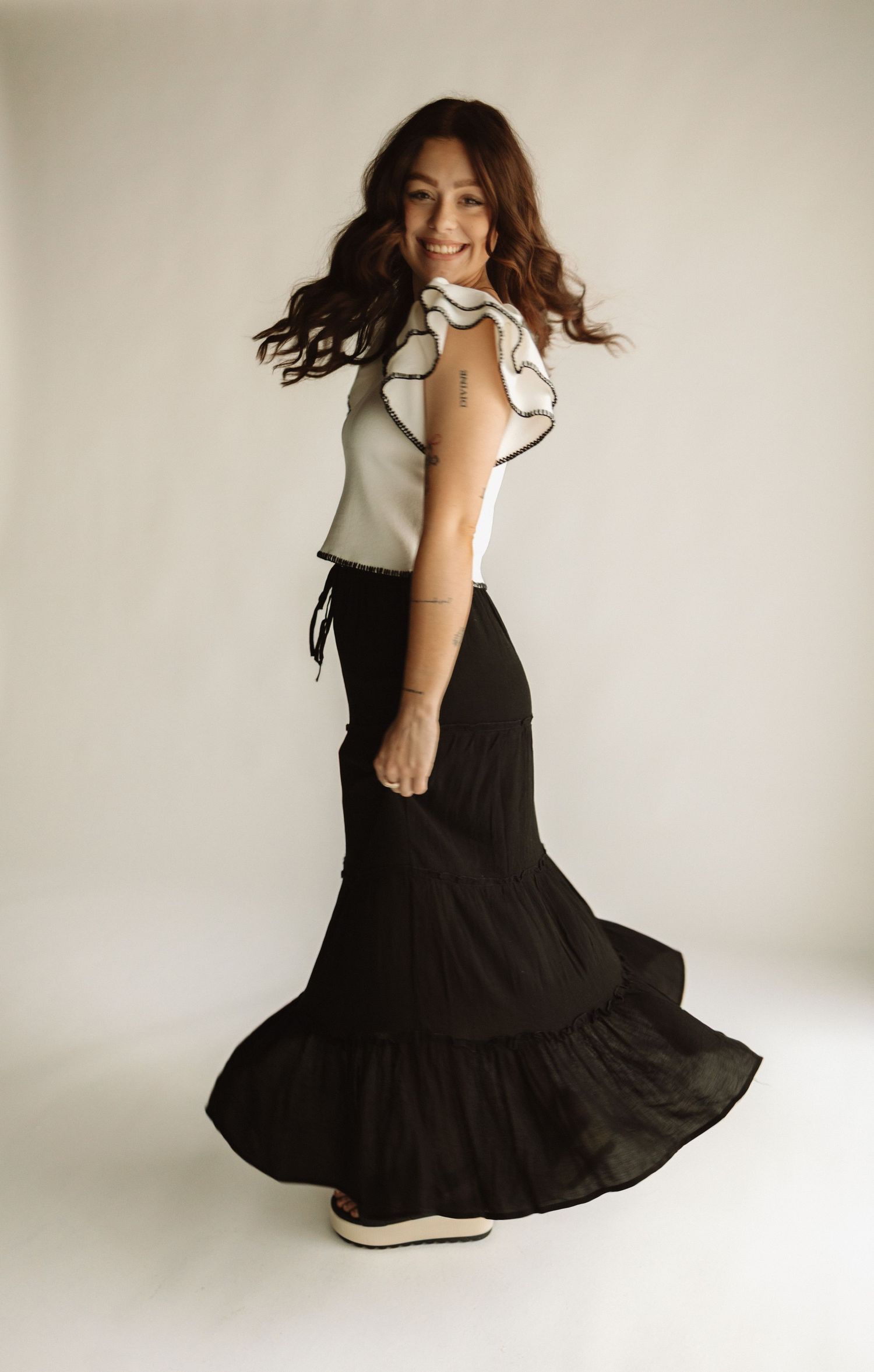 A person in a flowing black maxi skirt and white top twirls joyfully against a neutral backdrop.