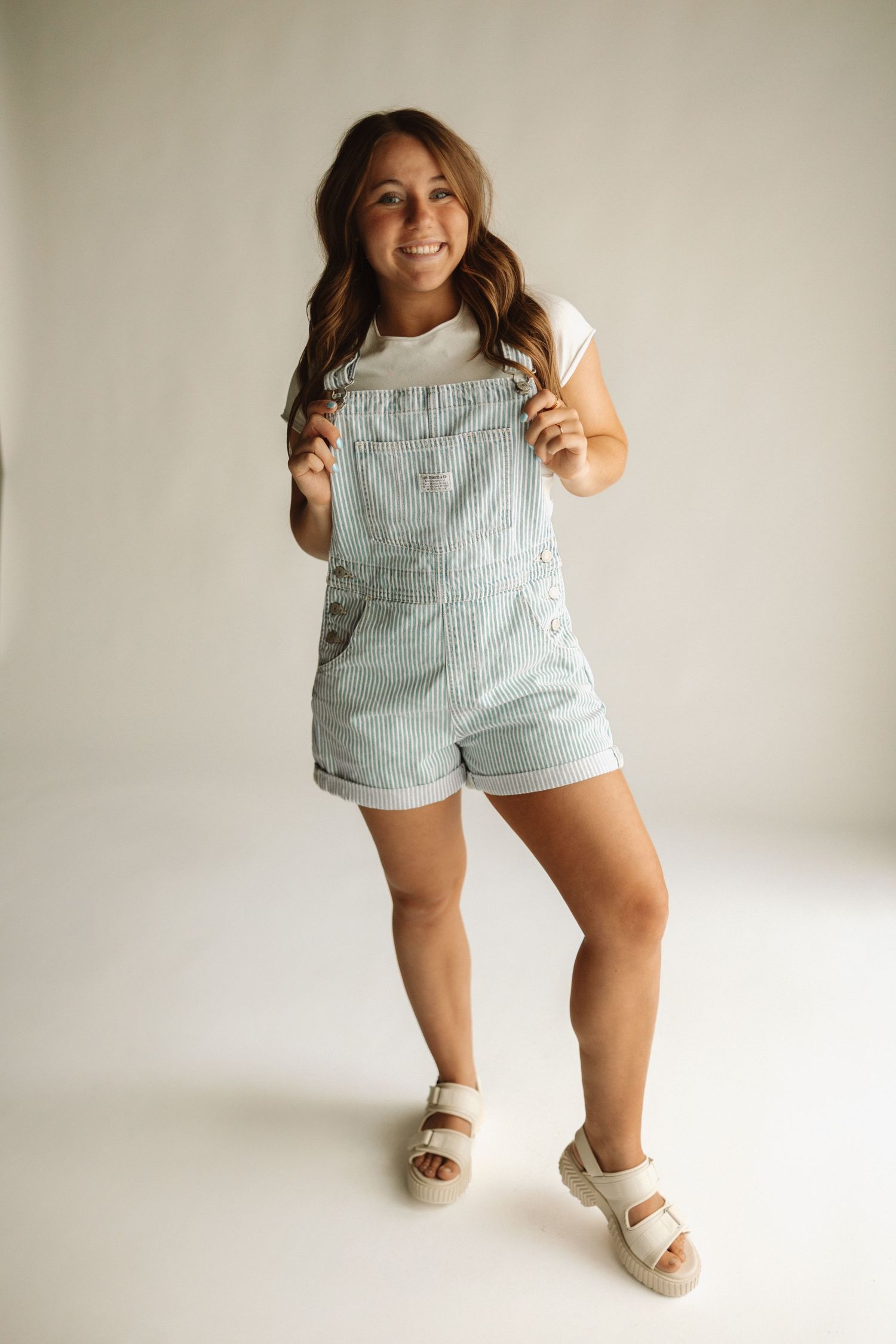 Person wearing light denim overall shorts and beige sandals posing against white wall in casual studio photoshoot.