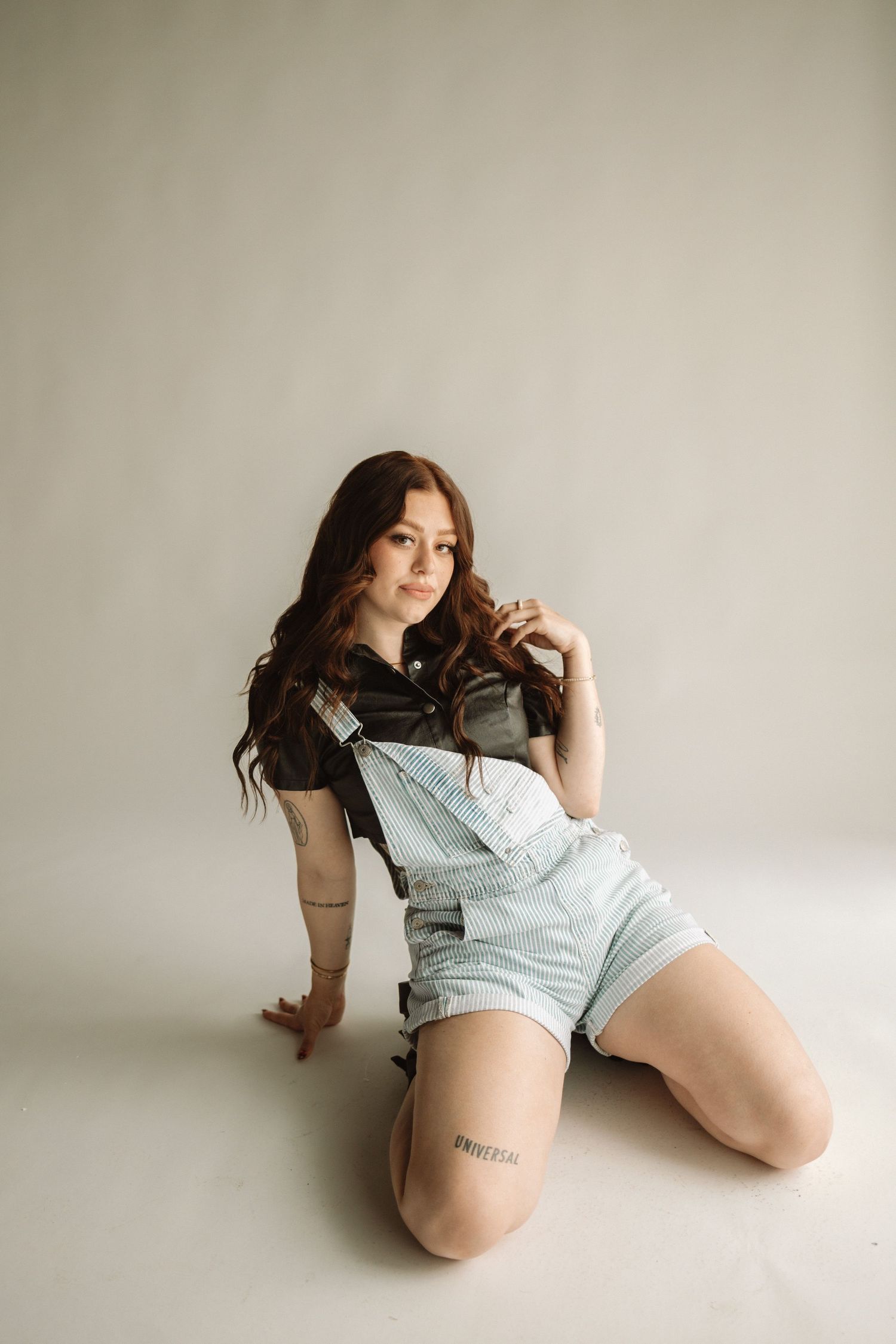 A person in light denim overalls and a black top poses casually on a light colored floor against a neutral wall.