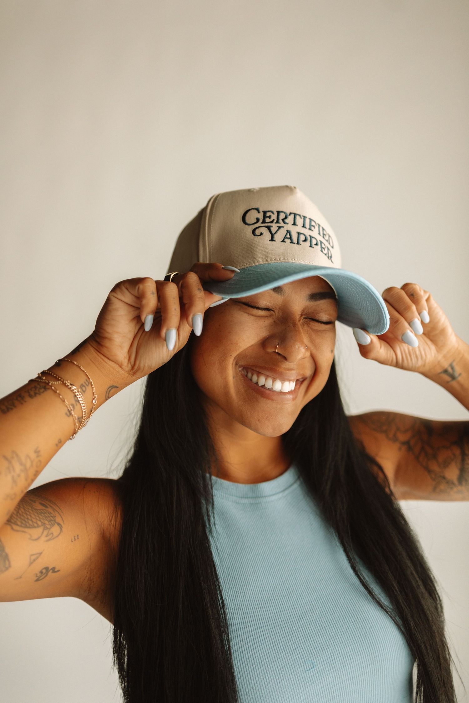 A joyful smile radiates beneath a tan baseball cap with 'Certified' text, paired with a light blue sleeveless top.
