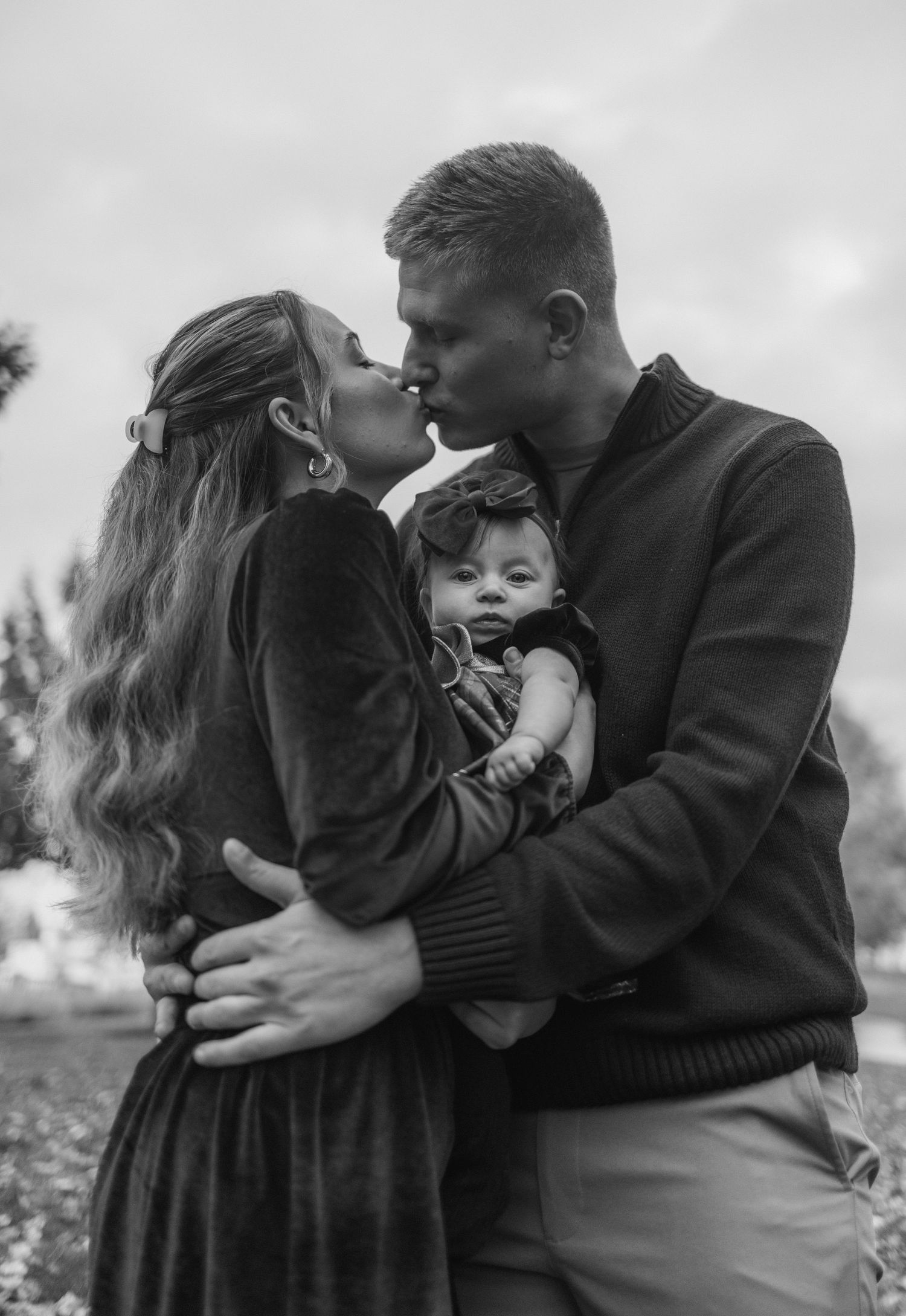 A tender black and white family portrait shows a couple sharing an intimate moment while holding their baby outdoors.