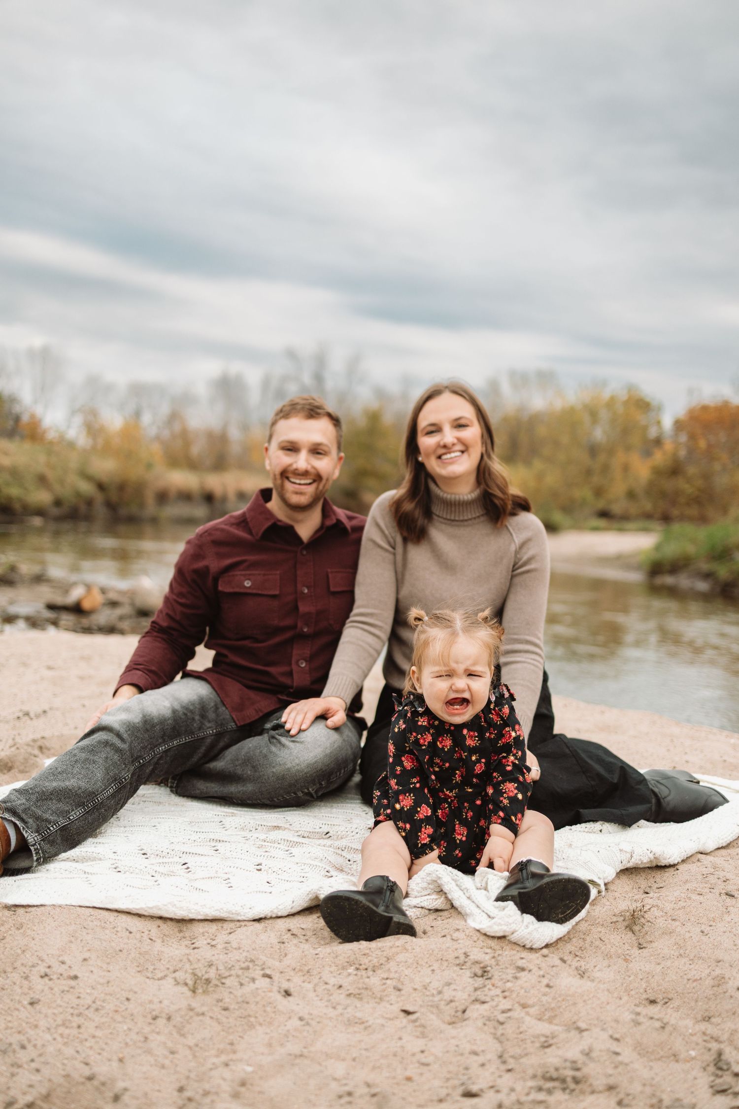 Family relaxing together on a blanket at a riverside beach during autumn with trees in the background.