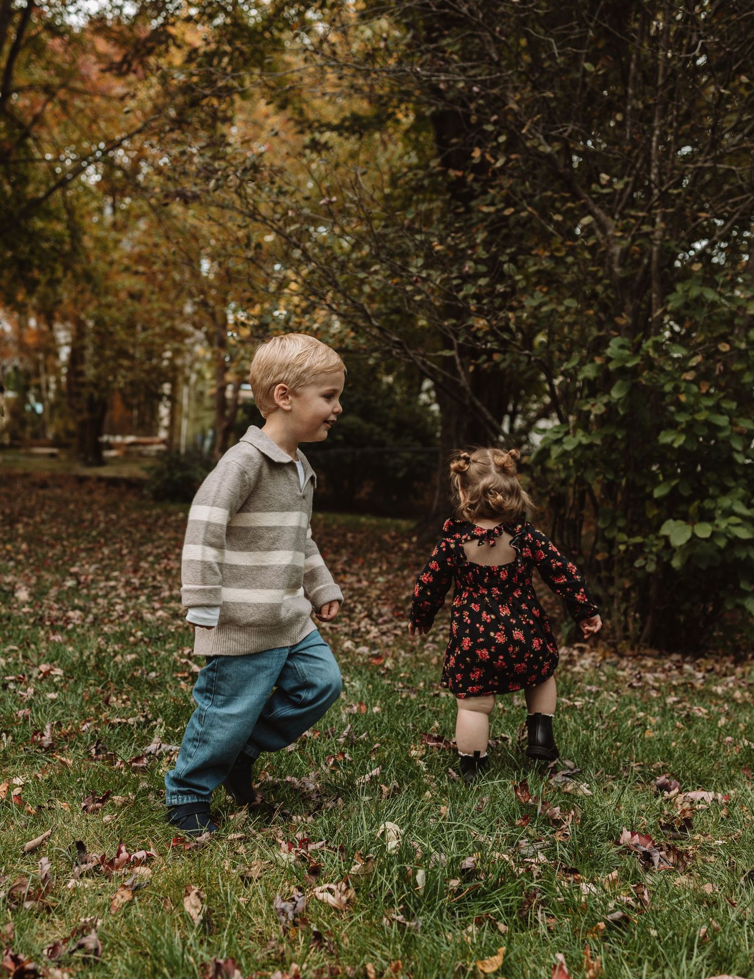 Two children explore a grassy area with fallen autumn leaves in a park setting.