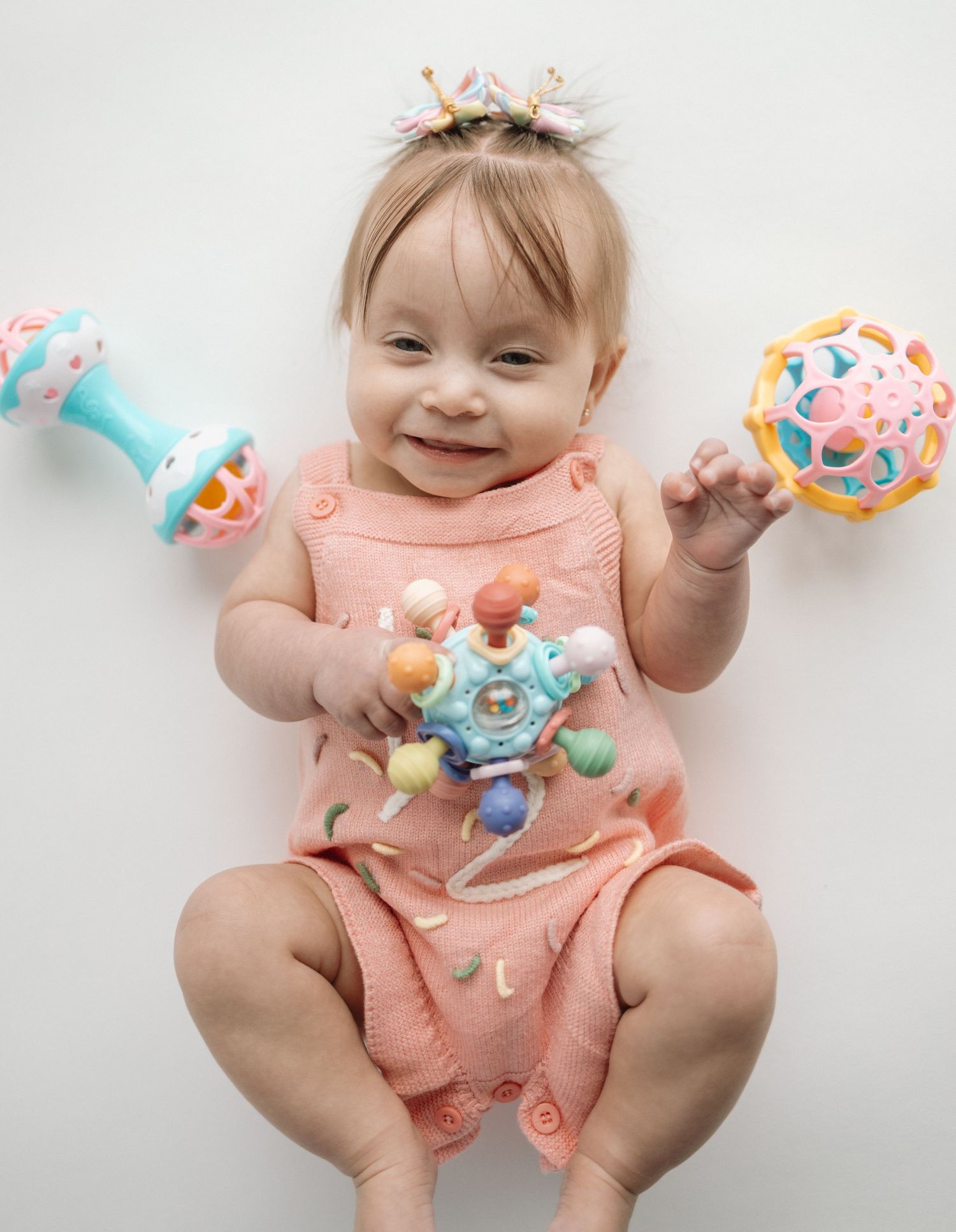 Baby in peach outfit playing with colorful teething toys while lying on white background.