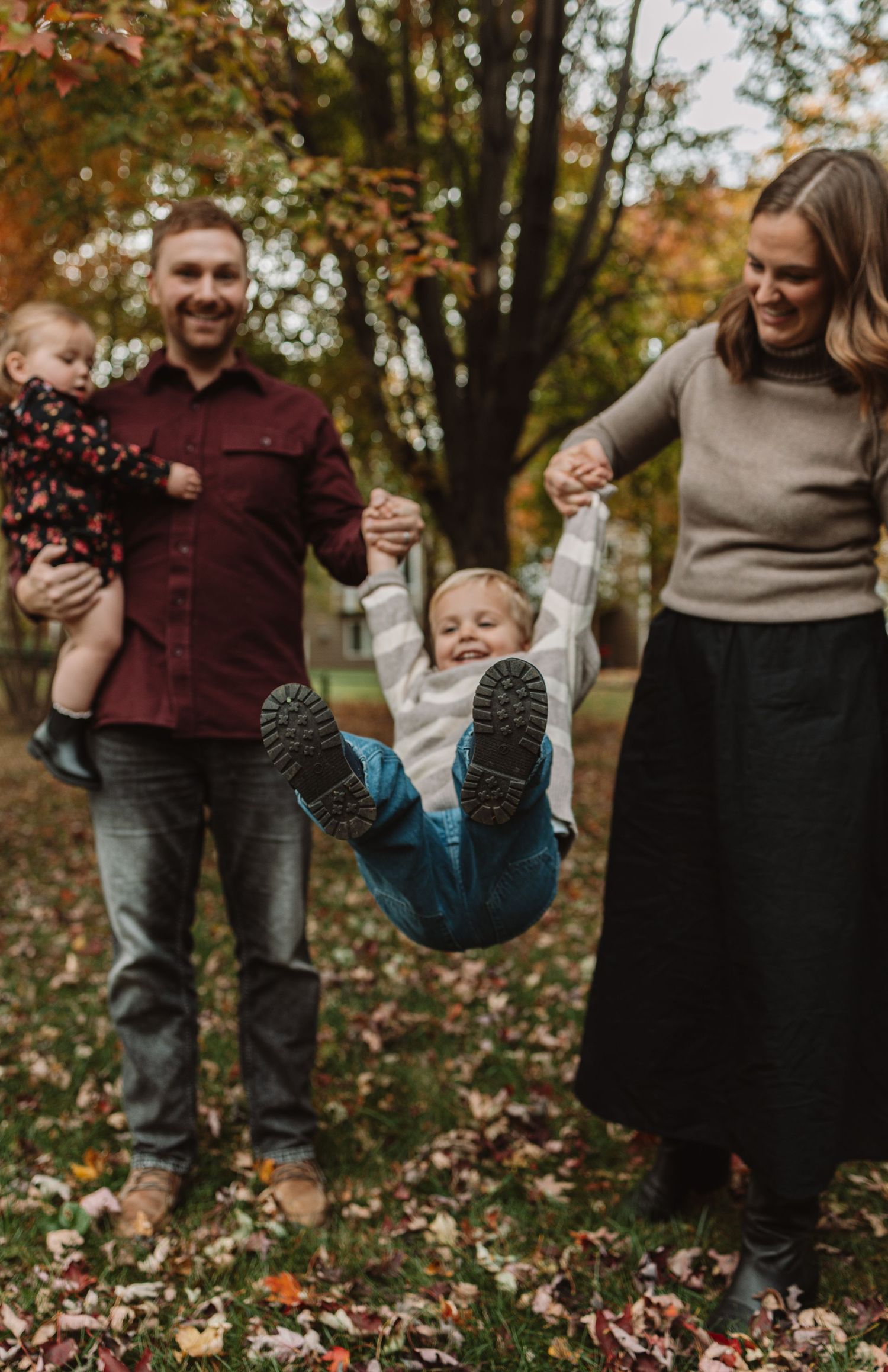Family plays and swings child in autumn leaves during outdoor photo session in park.