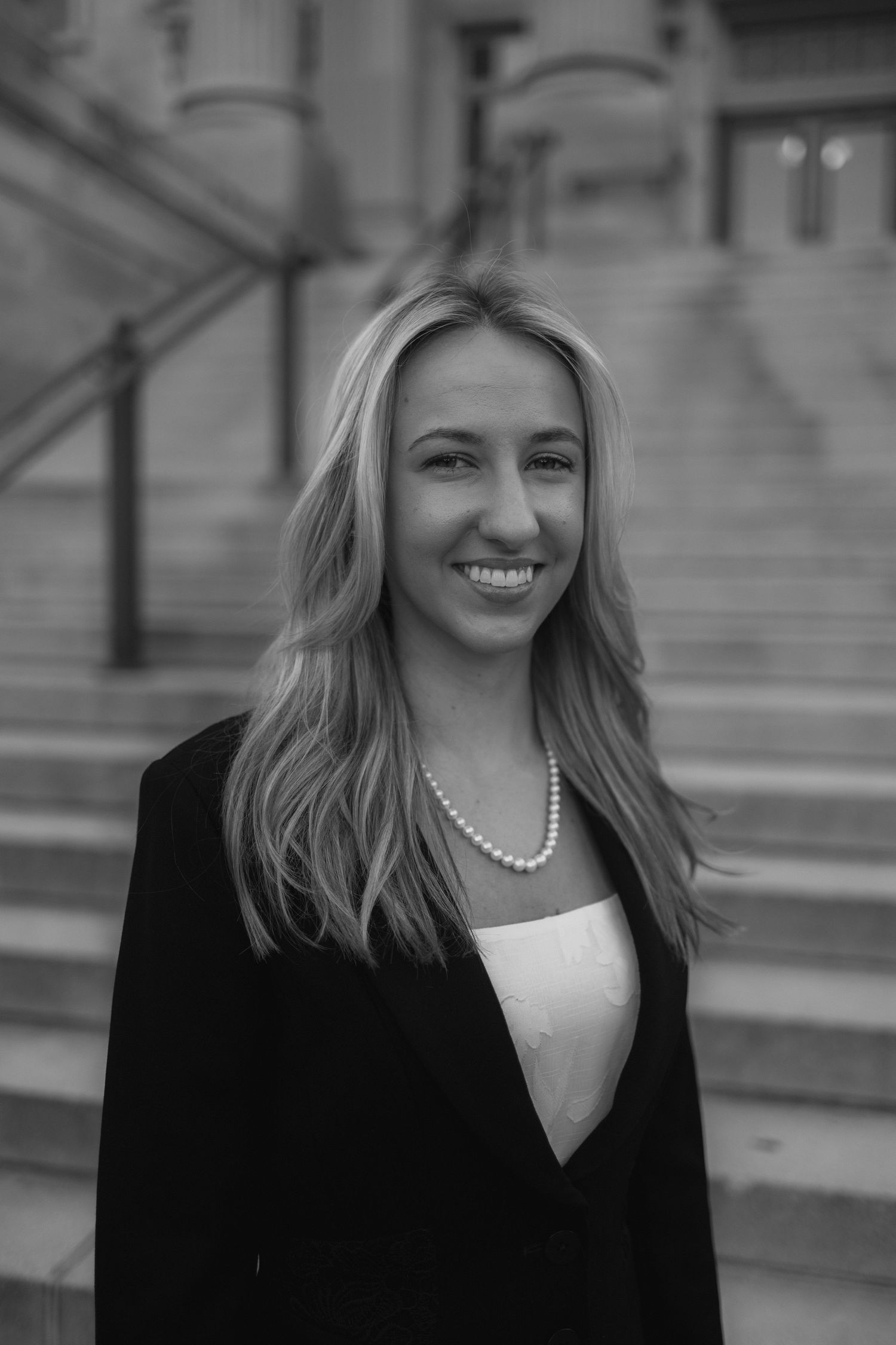 Professional portrait of someone in a black blazer and pearl necklace smiling on stone steps in black and white.