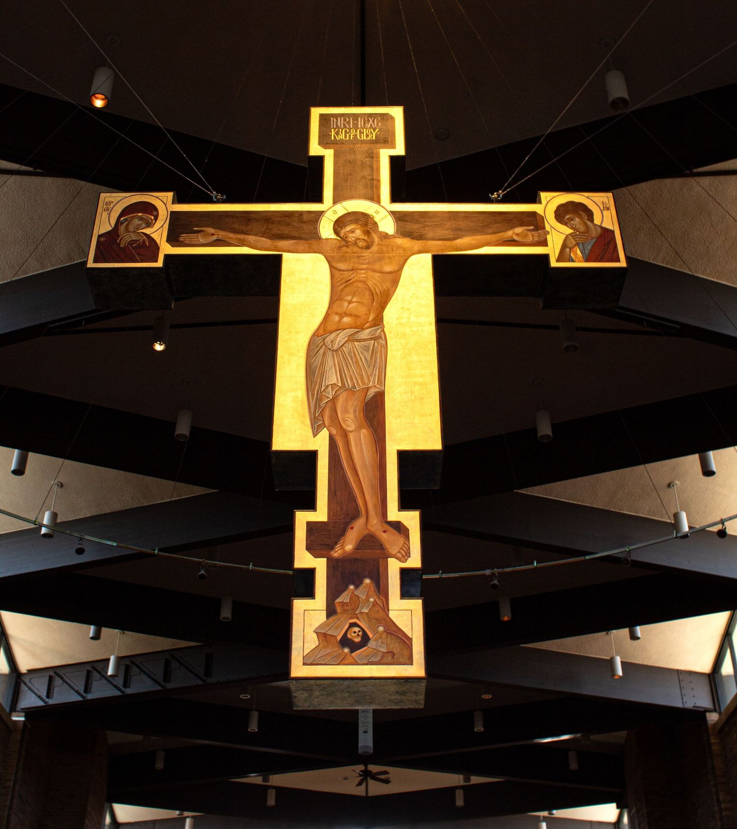 An illuminated crucifix hangs from a dark ceiling with geometric beams, creating a dramatic religious focal point.