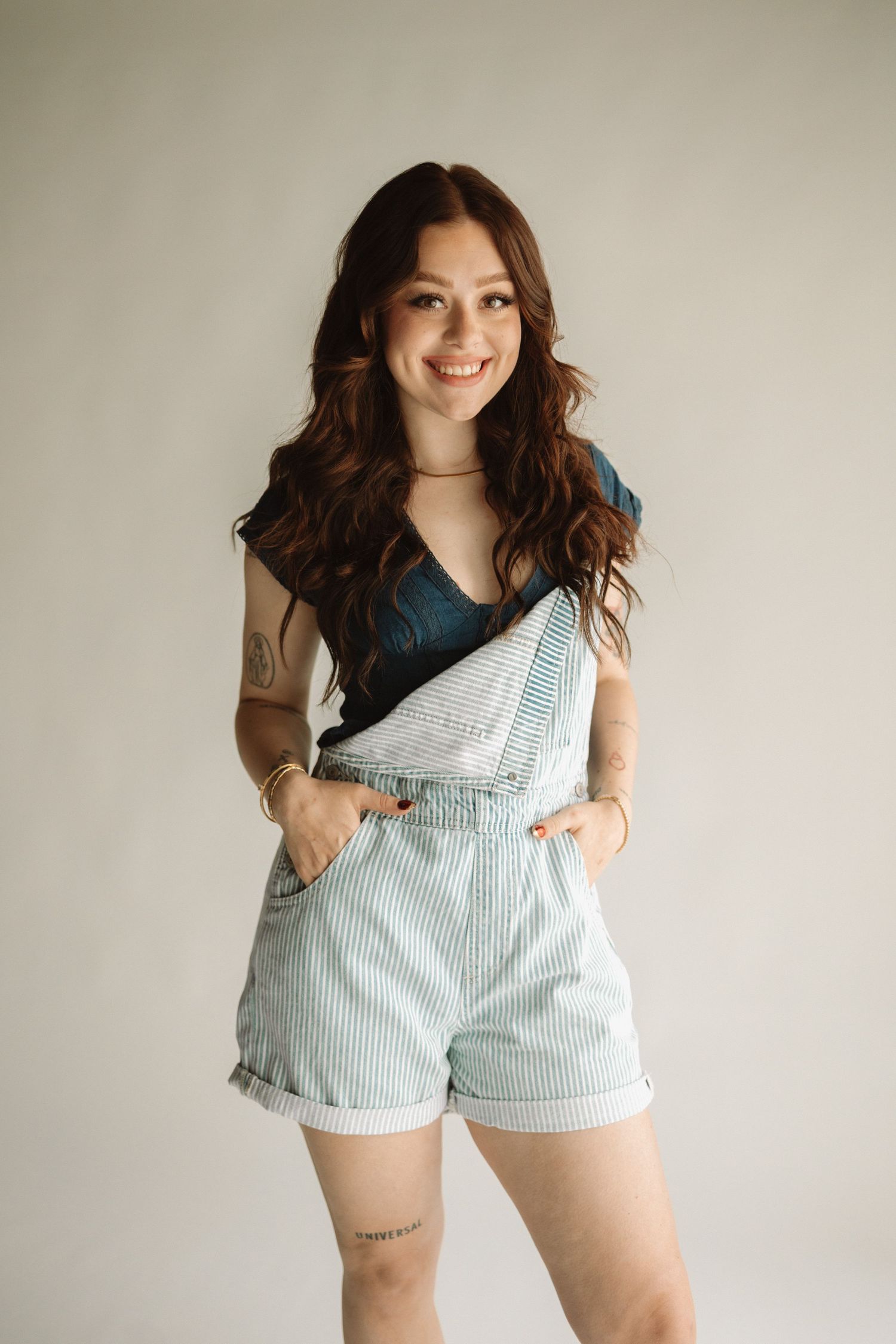 Casual summer outfit featuring light blue striped overall shorts paired with a dark teal top against neutral background.