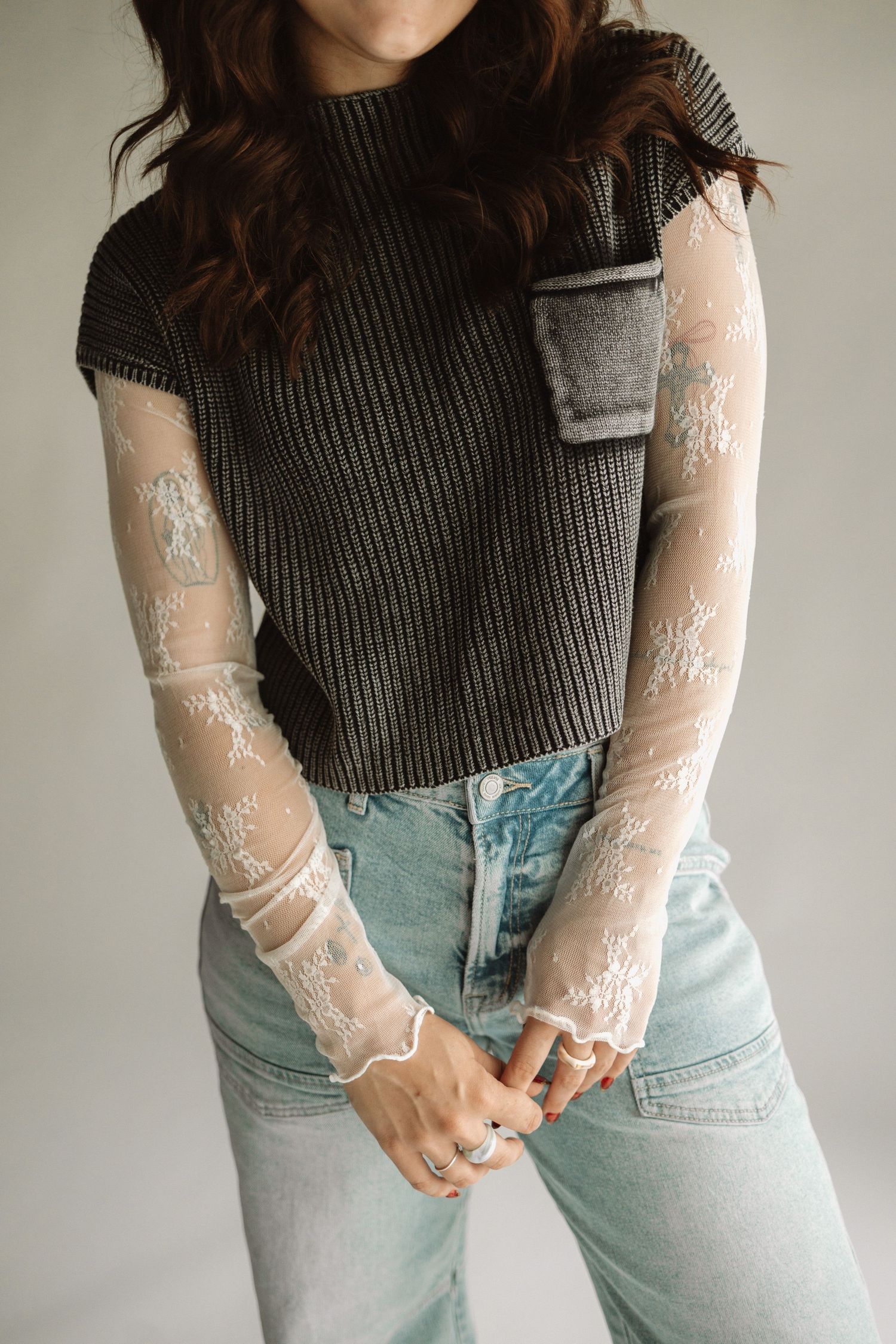 A person wearing a dark gray ribbed top with delicate lace sleeves and light wash high-waisted jeans stands against a wall.