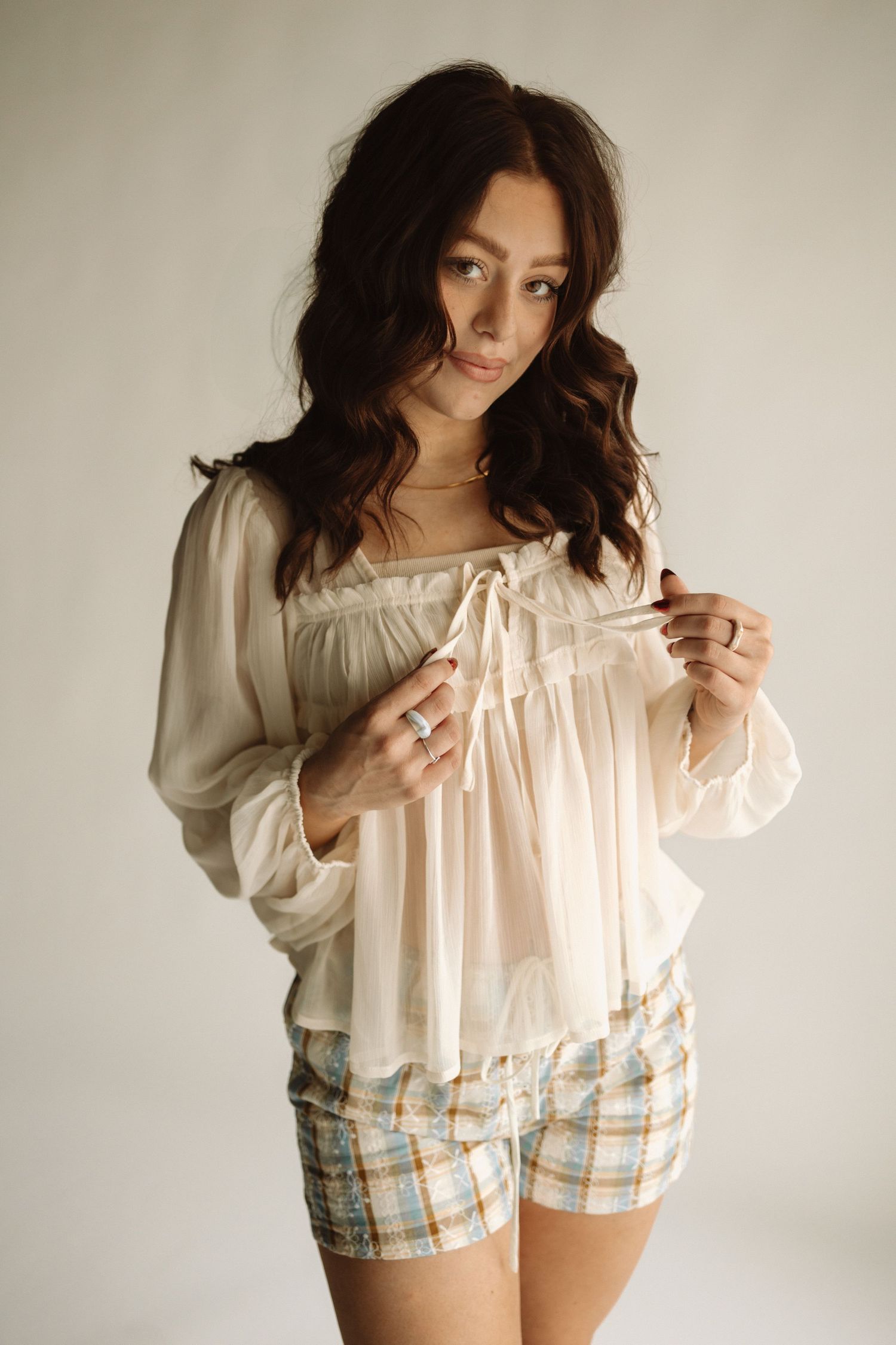 Woman in white blouse and plaid shorts poses against neutral background in a casual fashion photo series.