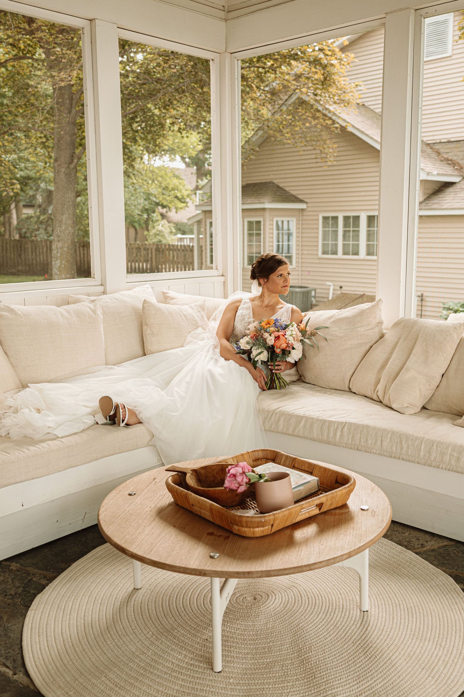 A white wedding dress and bouquet in a sunlit room with white sectional sofa and round coffee table on a screened porch.