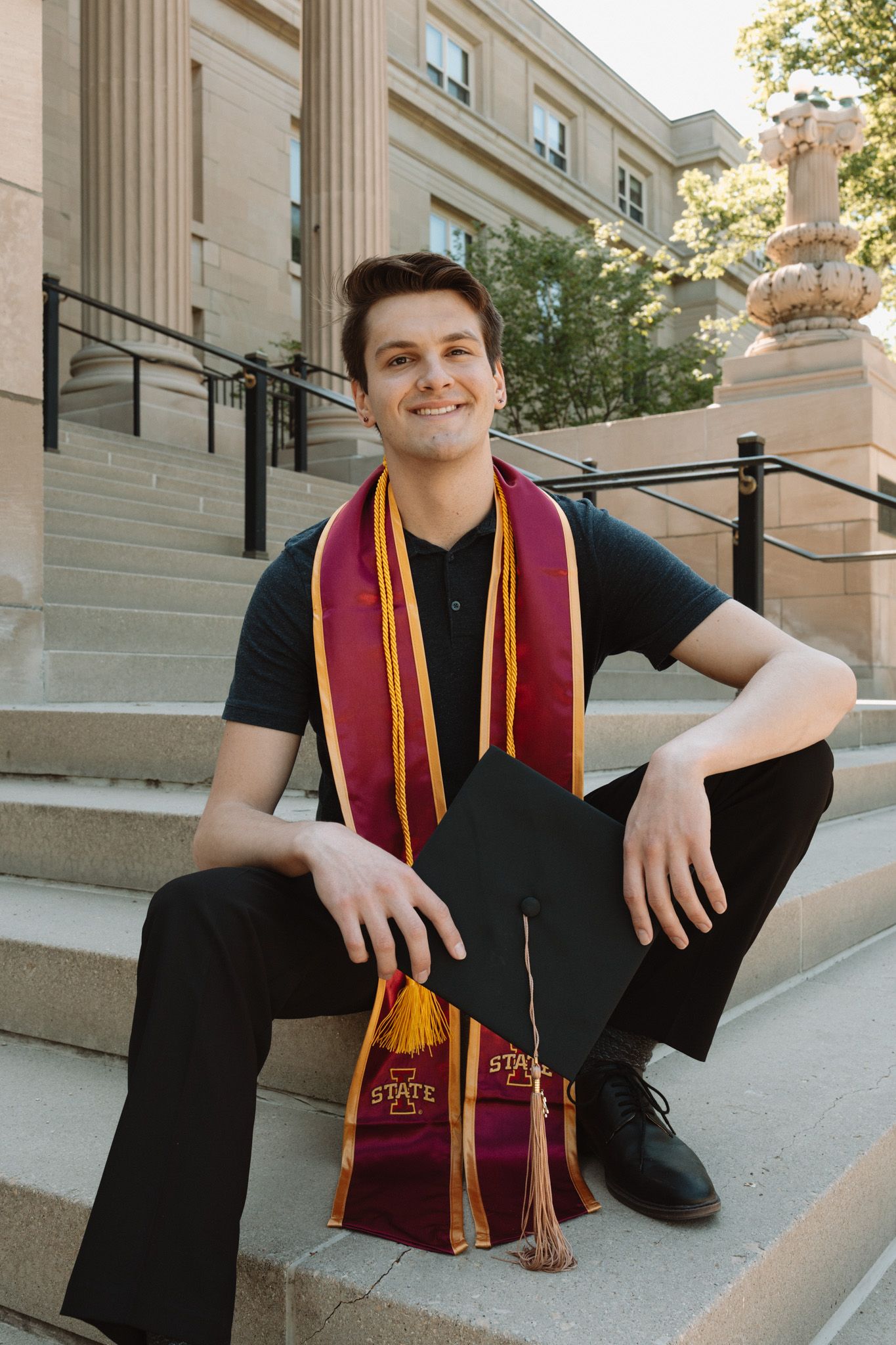 Graduate wearing academic regalia with burgundy and gold stole sits on concrete steps of campus building.