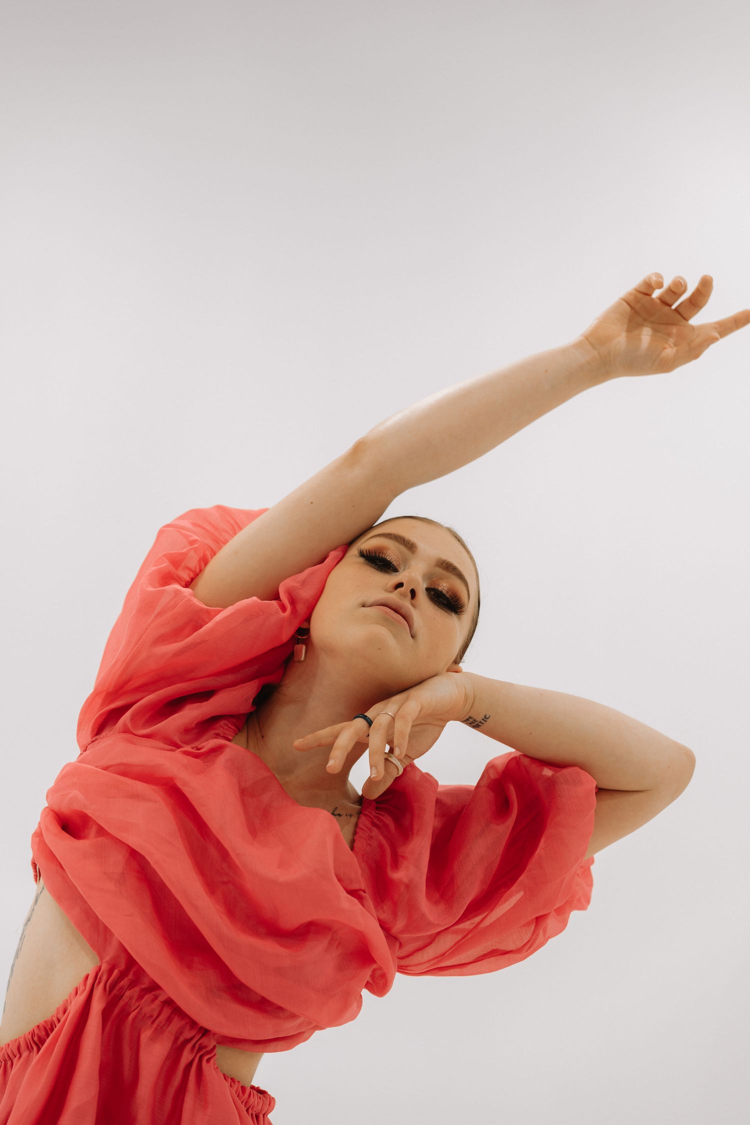 A figure in a flowing coral red dress poses gracefully with one arm raised against a light background.