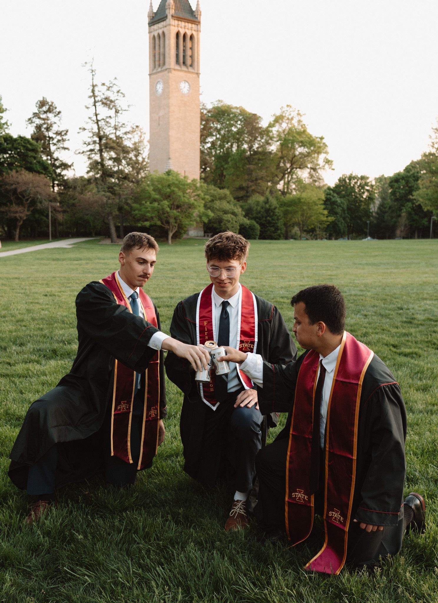 Three graduates in caps and gowns celebrating on campus lawn with historic tower in background.