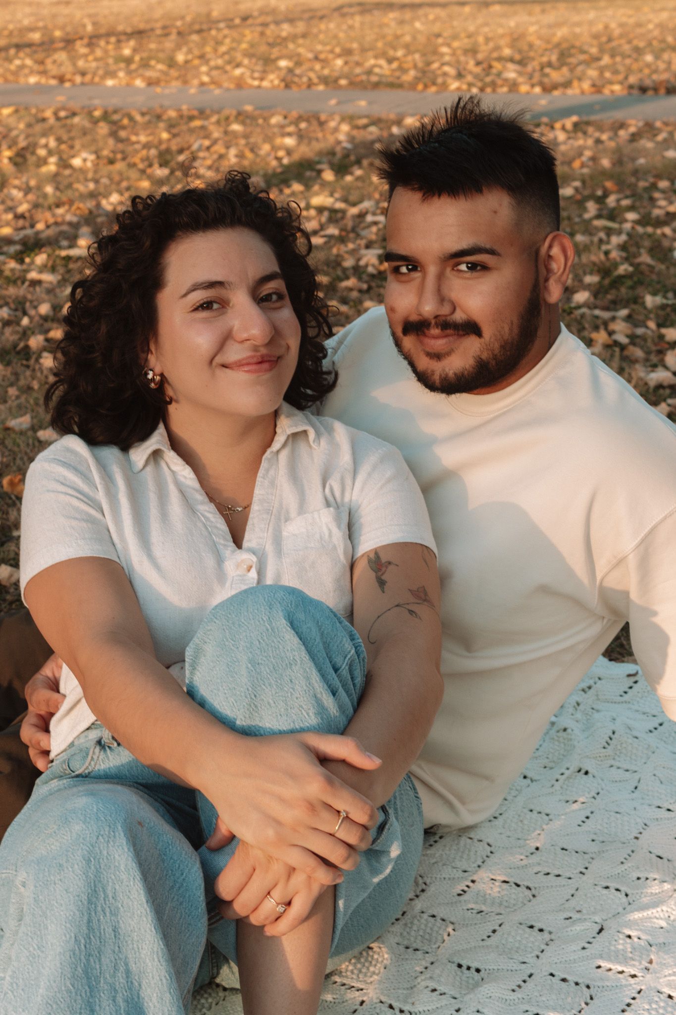 A couple in matching white shirts sits together on a blanket in an autumn outdoor setting.