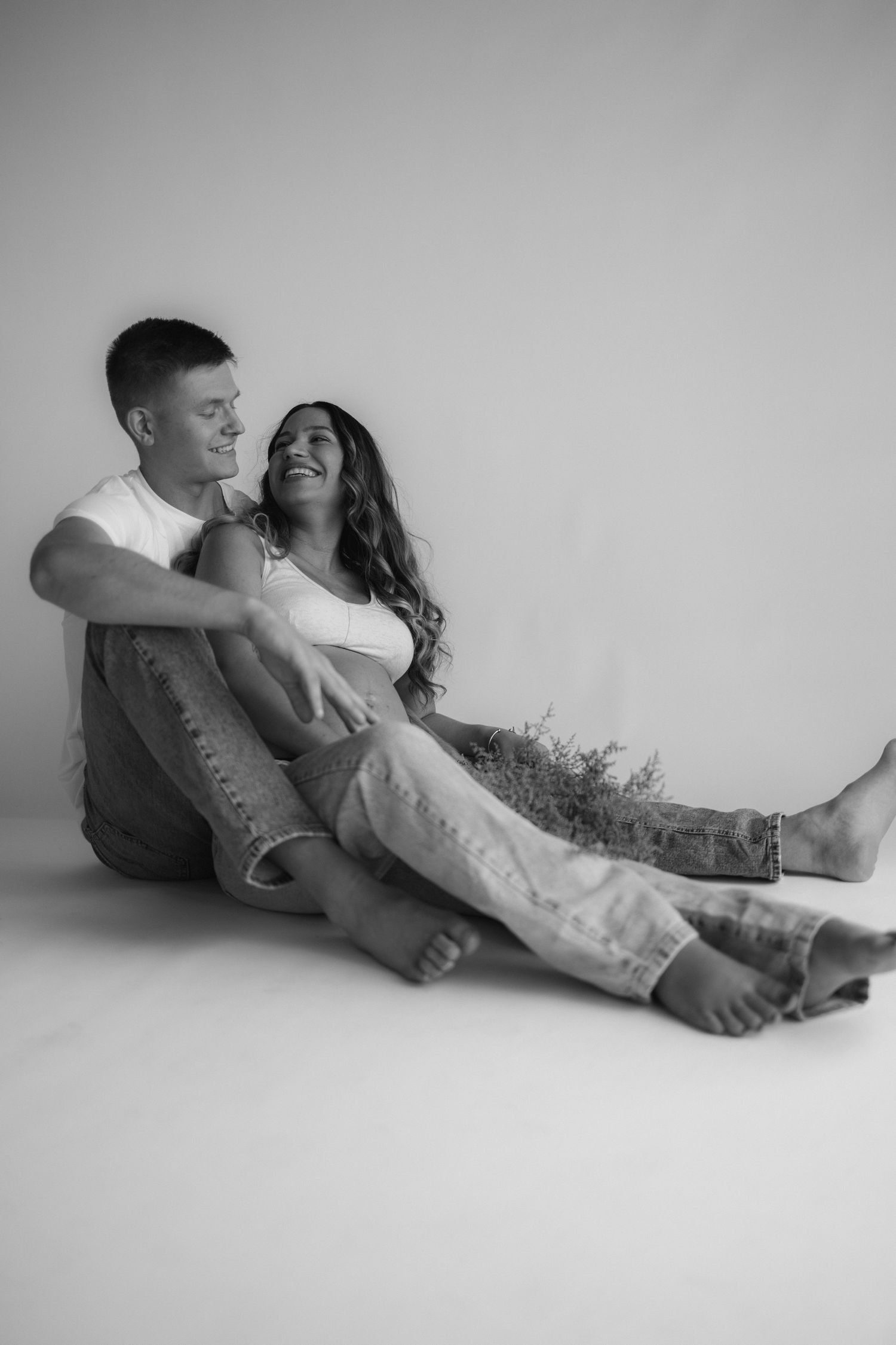 A couple sits together on a white floor laughing and embracing in a casual black and white portrait.