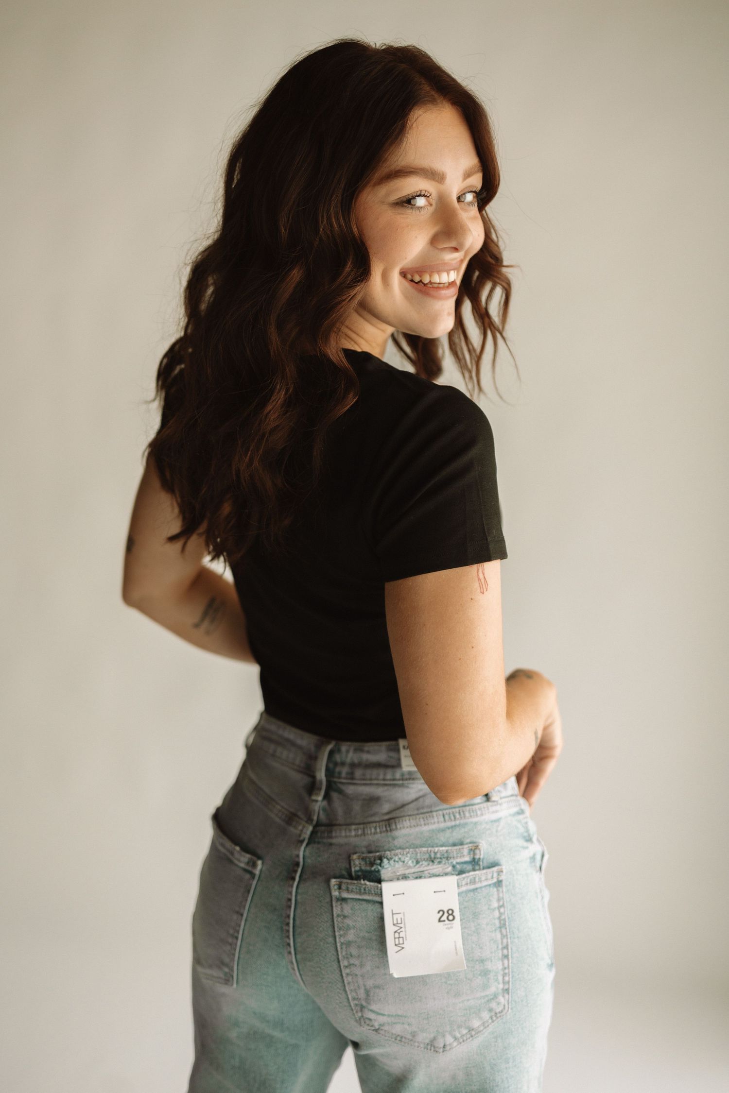A person with dark wavy hair poses in a black t-shirt and high-waisted jeans while smiling and looking over their shoulder.