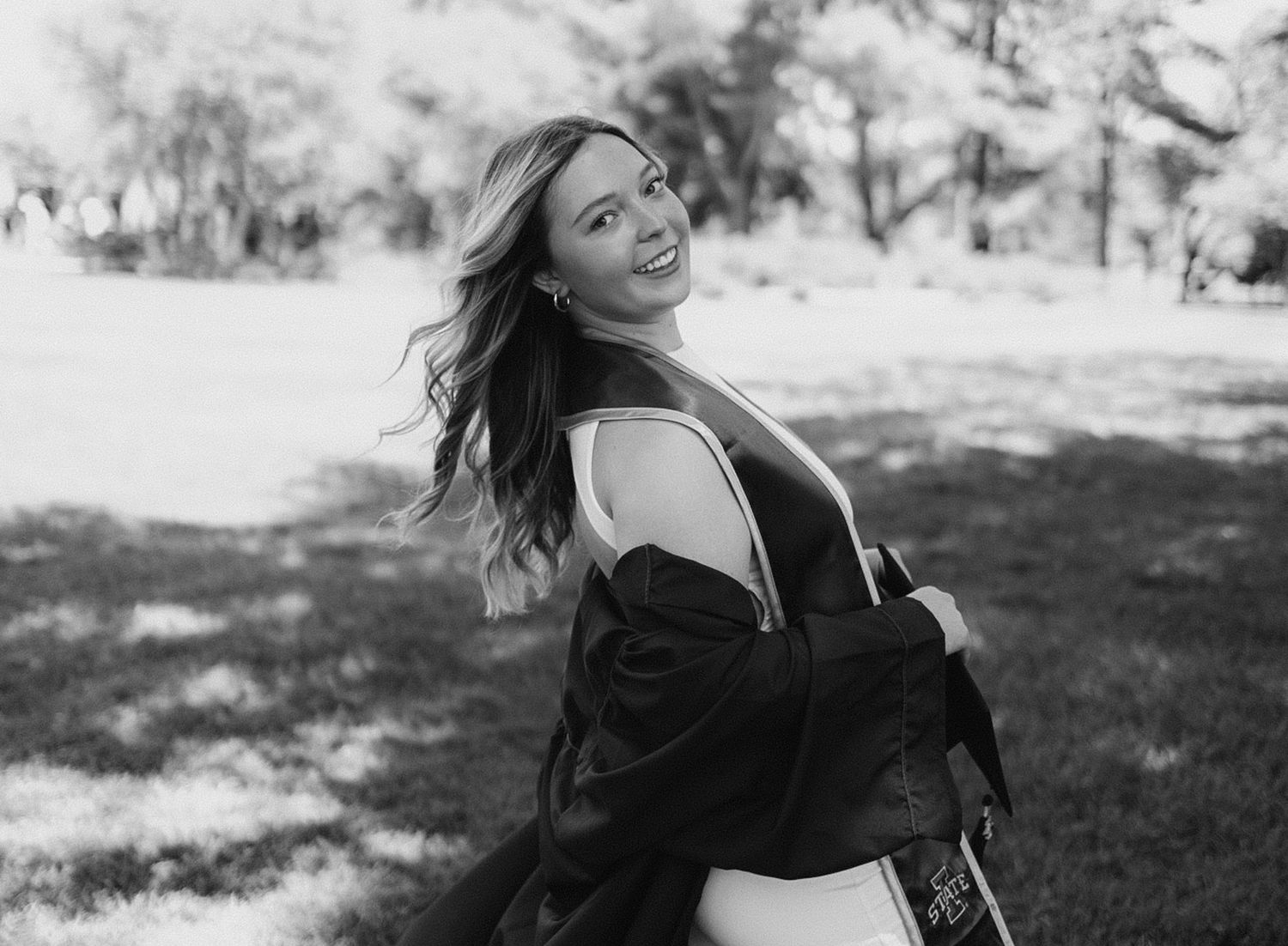 Graduate in academic regalia poses outdoors with flowing hair and joyful expression.