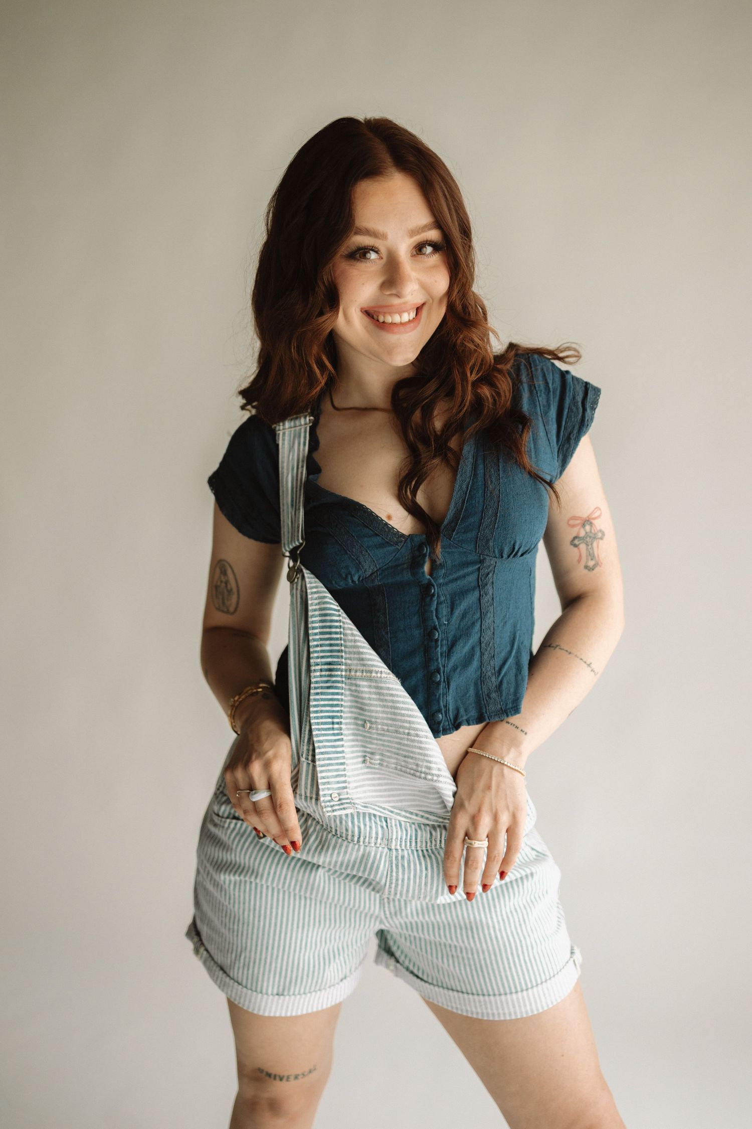 Smiling individual in casual striped overalls and dark shirt poses against neutral wall showing arm tattoos.