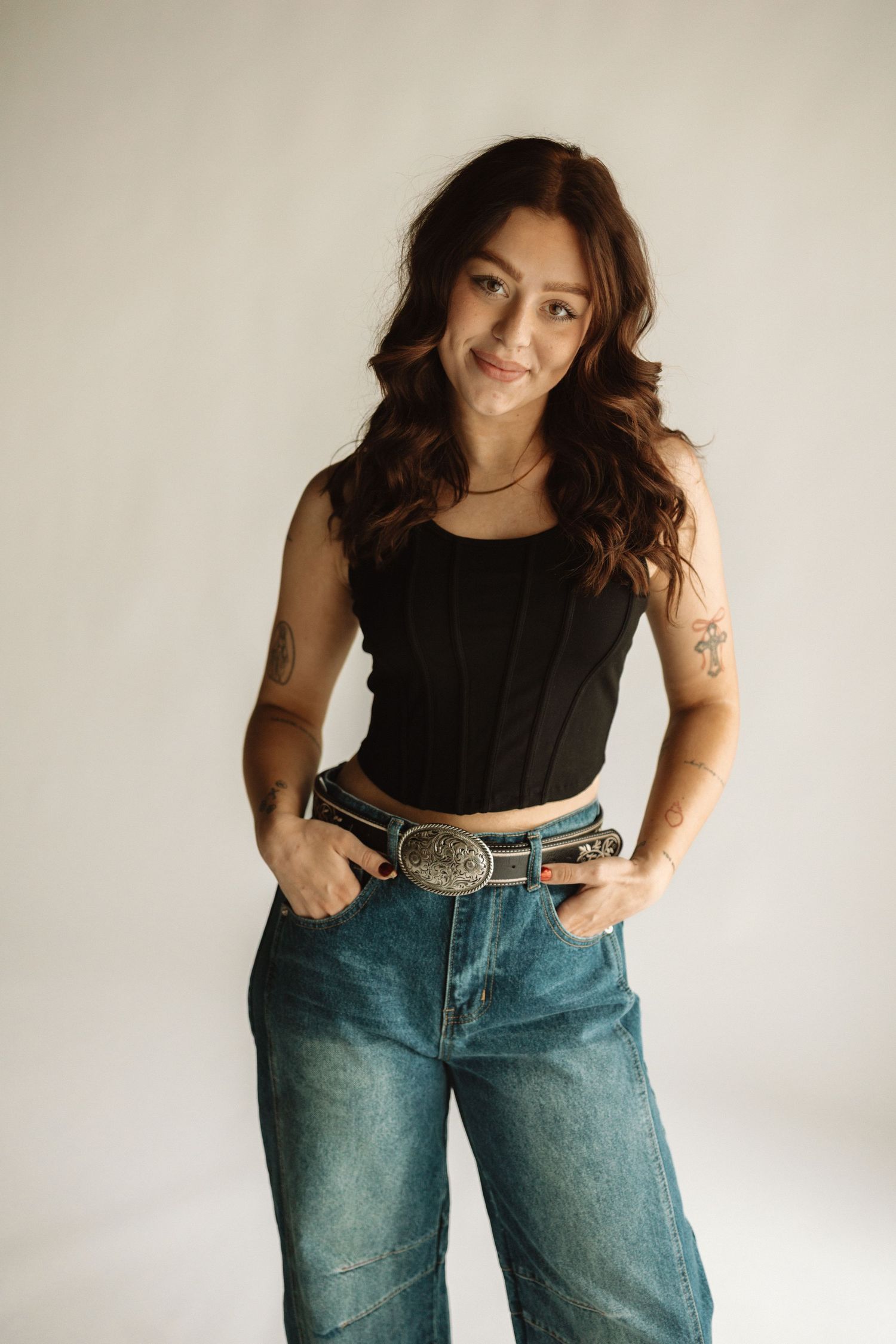 A person poses in casual attire wearing a black sleeveless top, blue jeans, and a statement belt against a white wall.