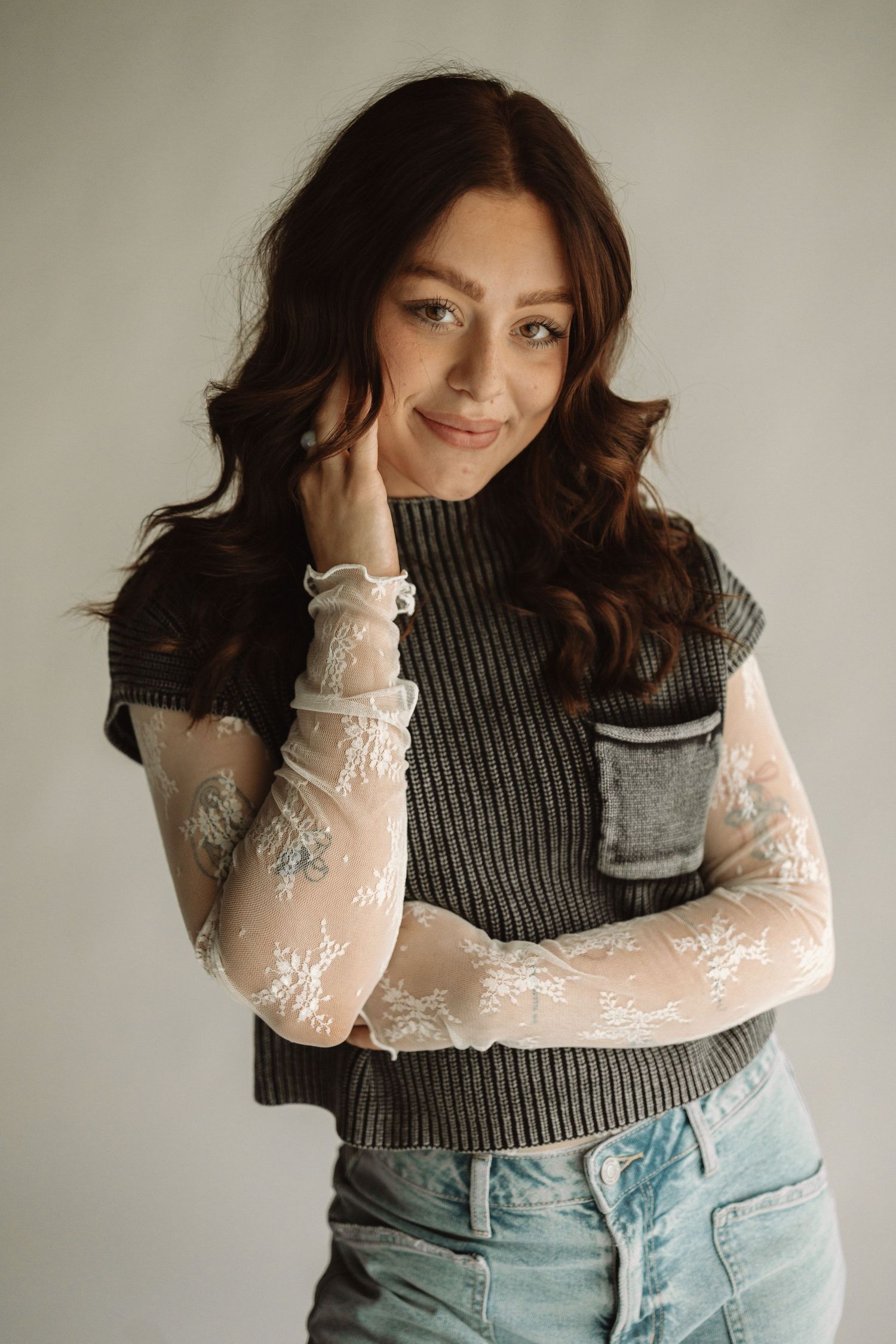 Portrait in casual attire featuring grey ribbed top with delicate lace sleeves and denim against light backdrop.