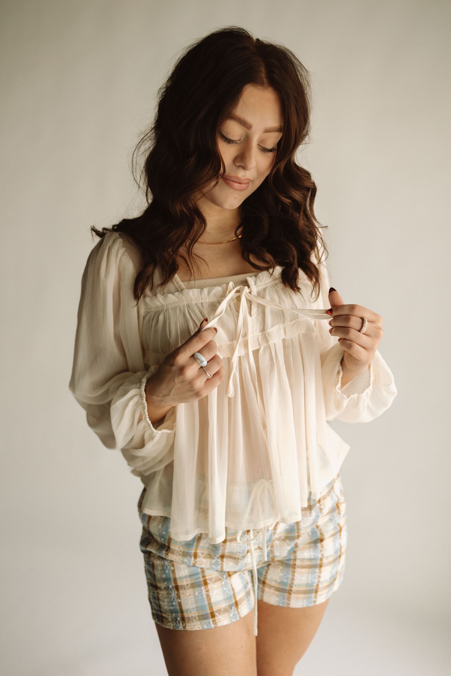 Woman in romantic cream peasant blouse and plaid shorts poses against light background in soft studio lighting.