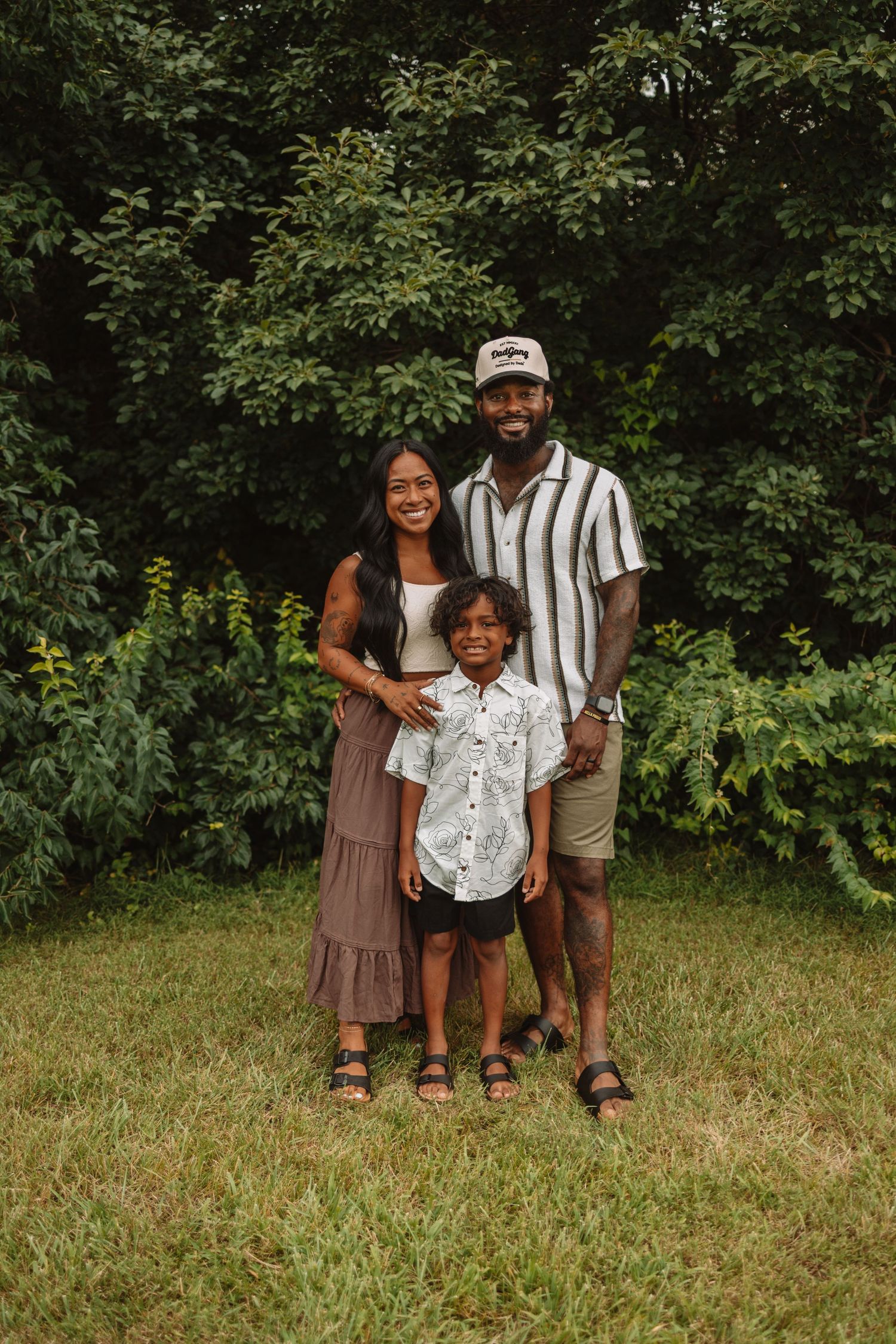A family poses together outdoors in coordinated summer outfits against a leafy green background.