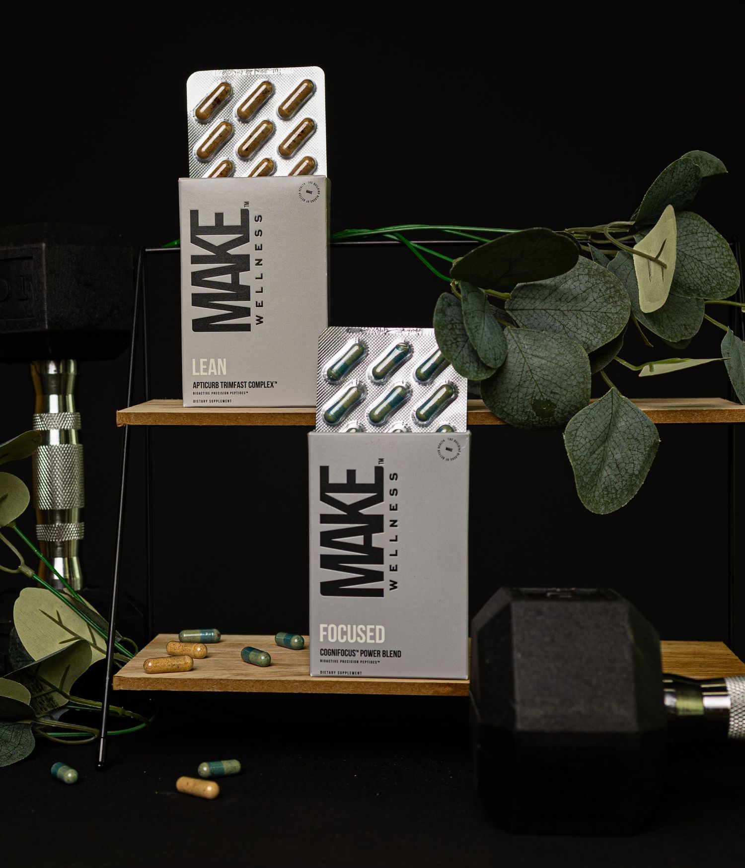 Two white MAKE supplement boxes displayed on wooden shelves with green eucalyptus leaves and black background.