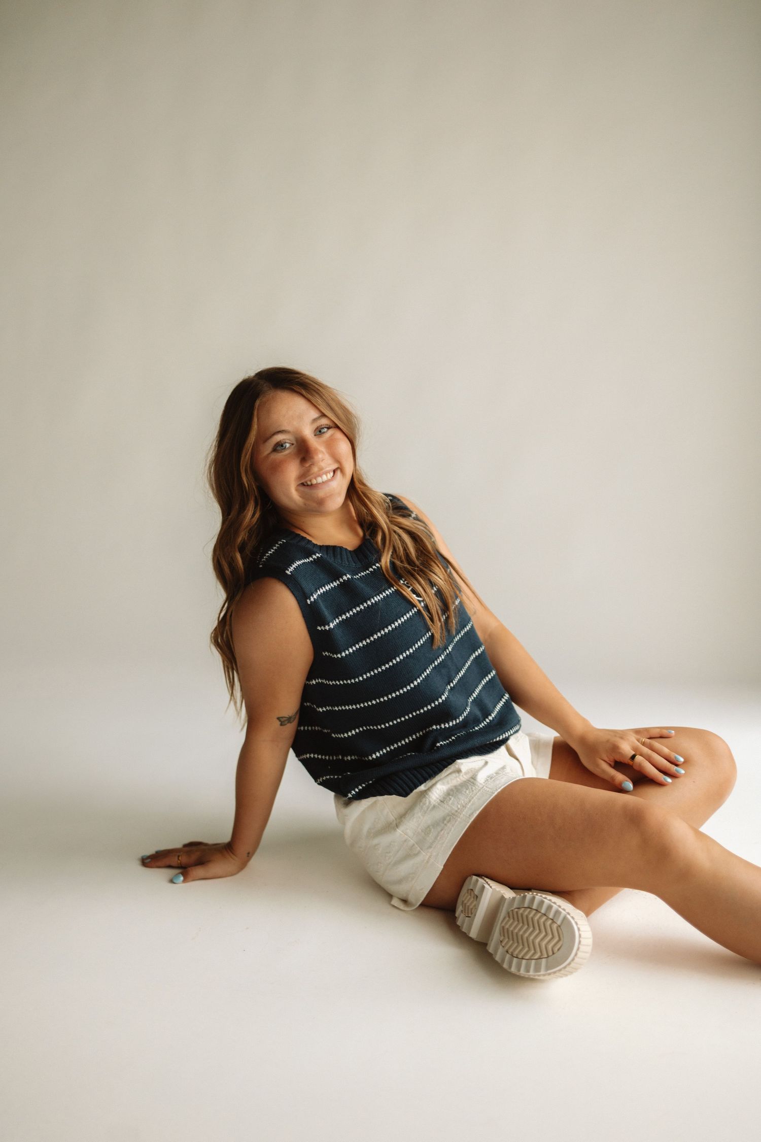 A person in a navy striped tank top and white shorts sits casually on a white floor against a neutral backdrop.