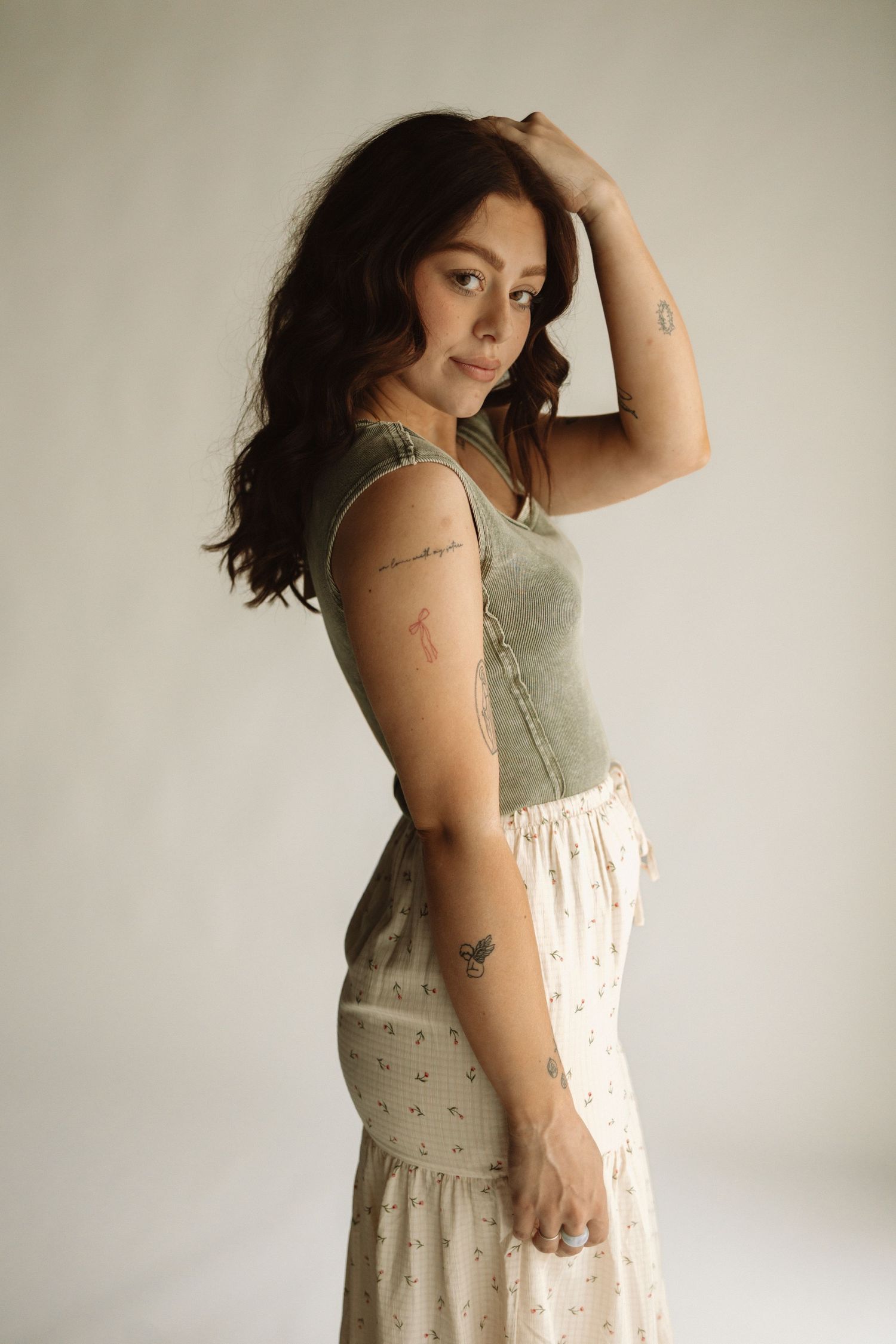 A woman in a sage green tank top and floral skirt poses against a light backdrop in a dreamy portrait.