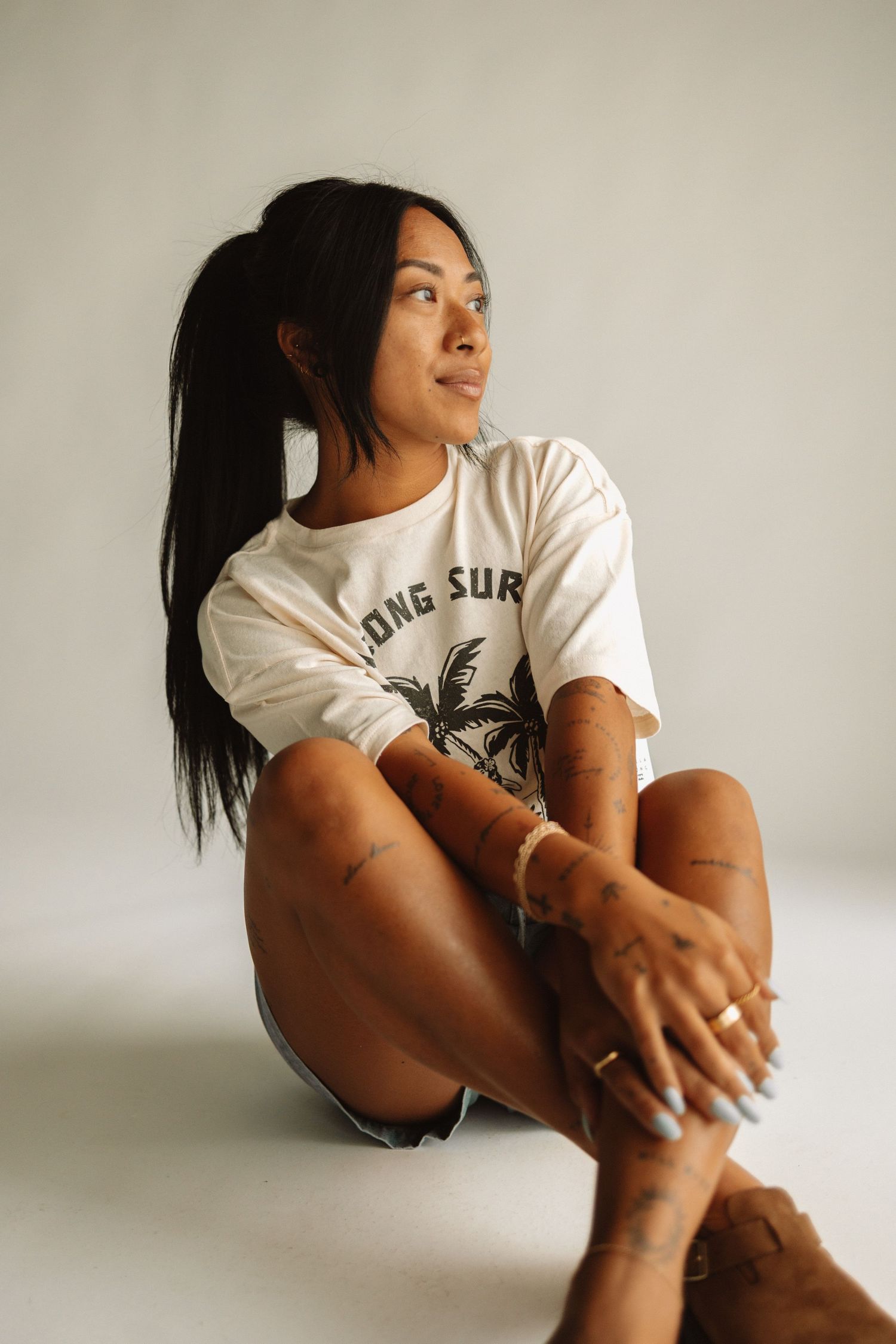 Someone sits on the floor wearing a casual white graphic t-shirt with long dark hair looking to the side against a light background.