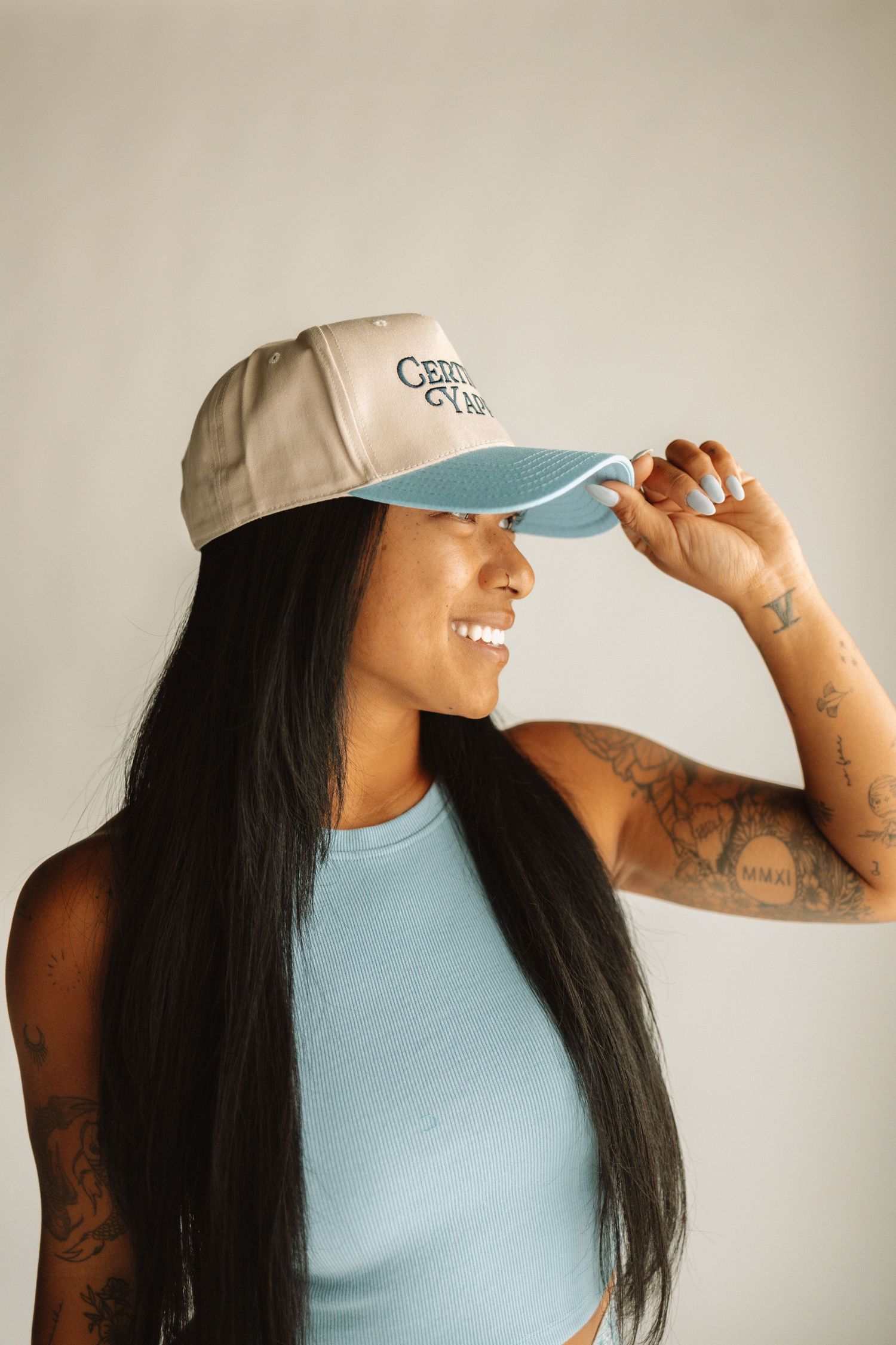 Person wearing a light blue tank top and beige baseball cap with long dark hair and visible arm tattoos.