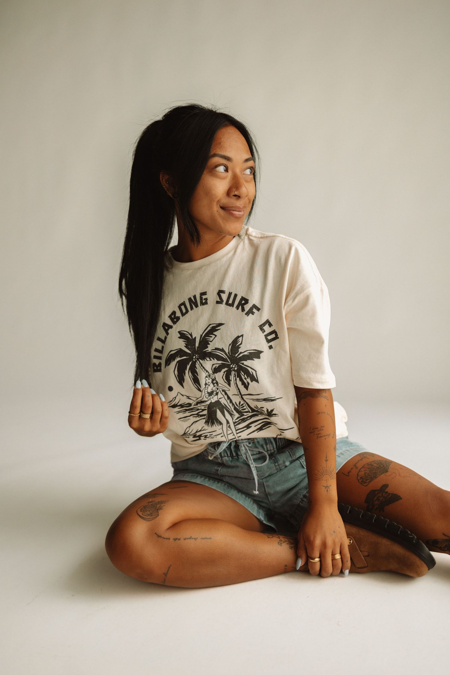Person in casual summer attire with Ragtong Surfer graphic tee and denim shorts lounging on white floor with natural lighting.