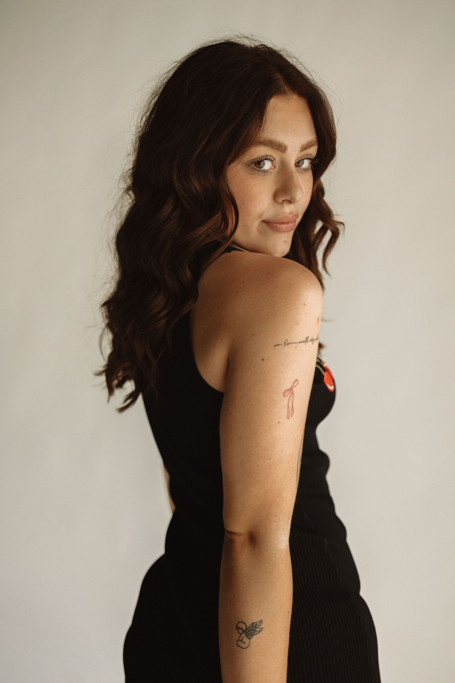 Portrait of someone in a black sleeveless top with dark wavy hair and small tattoos, looking over their shoulder.