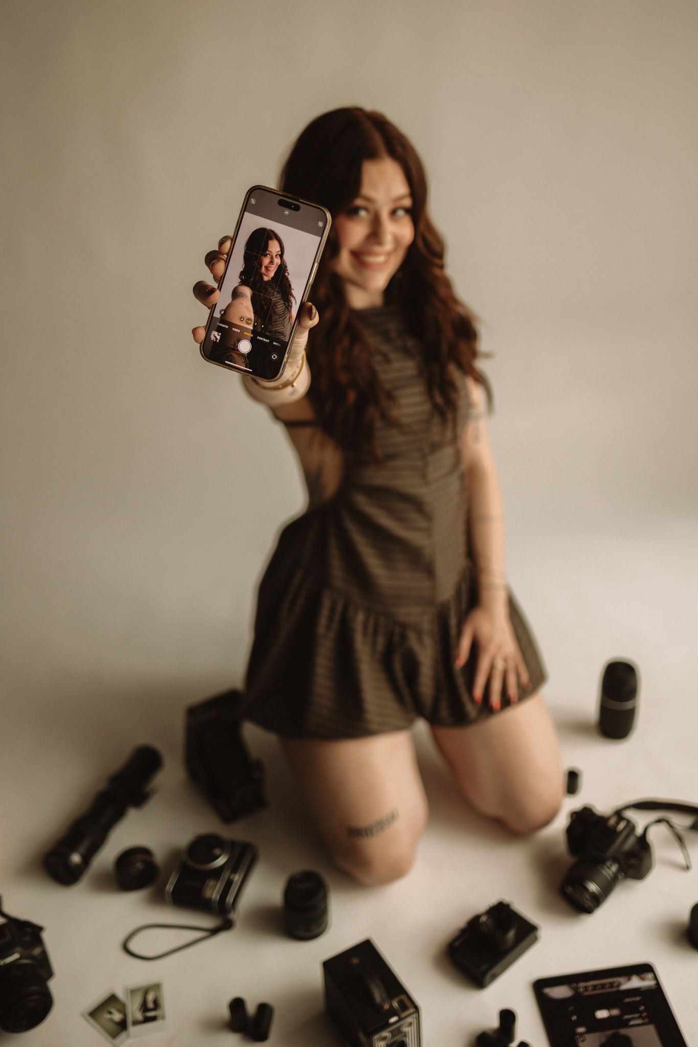 A photographer displays a selfie on their phone while kneeling among various camera equipment on the floor.