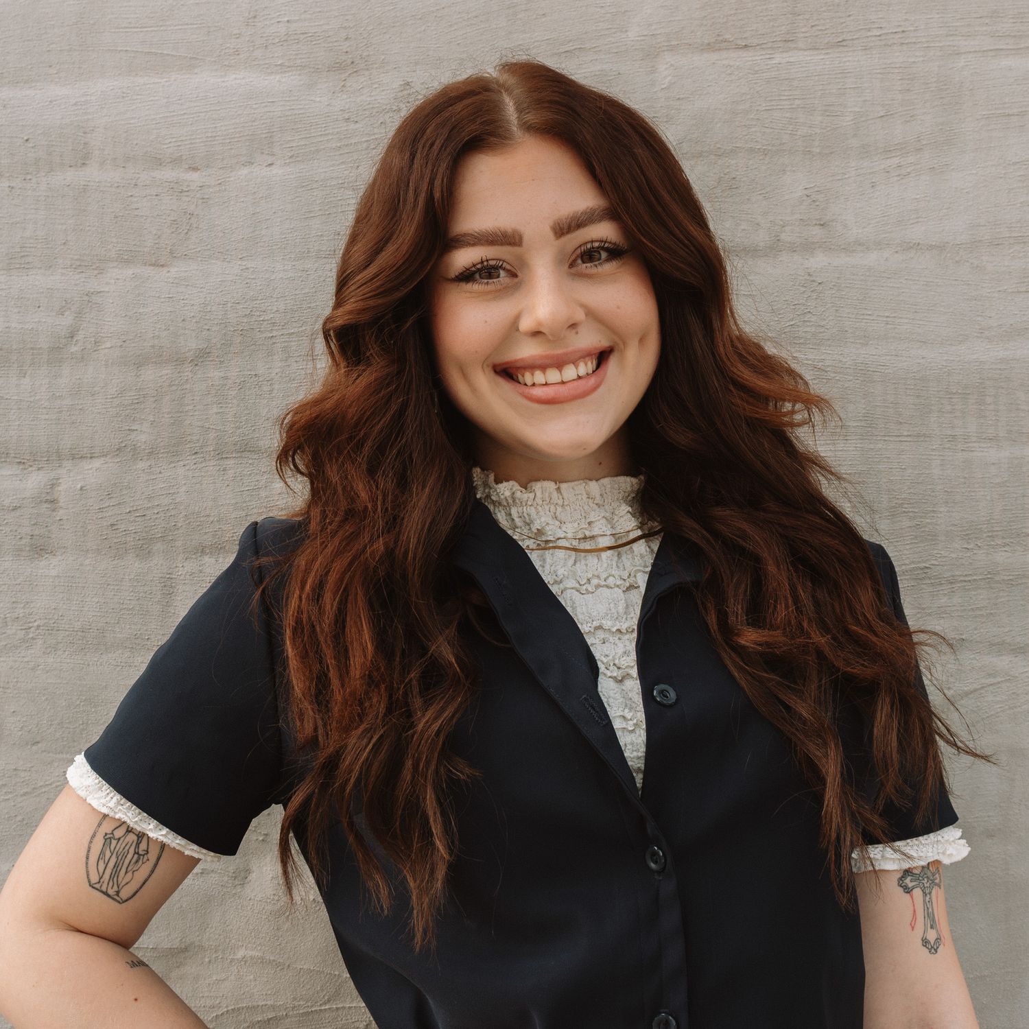 Person with long wavy brown hair wearing a black vest and white turtleneck smiles warmly against light background.