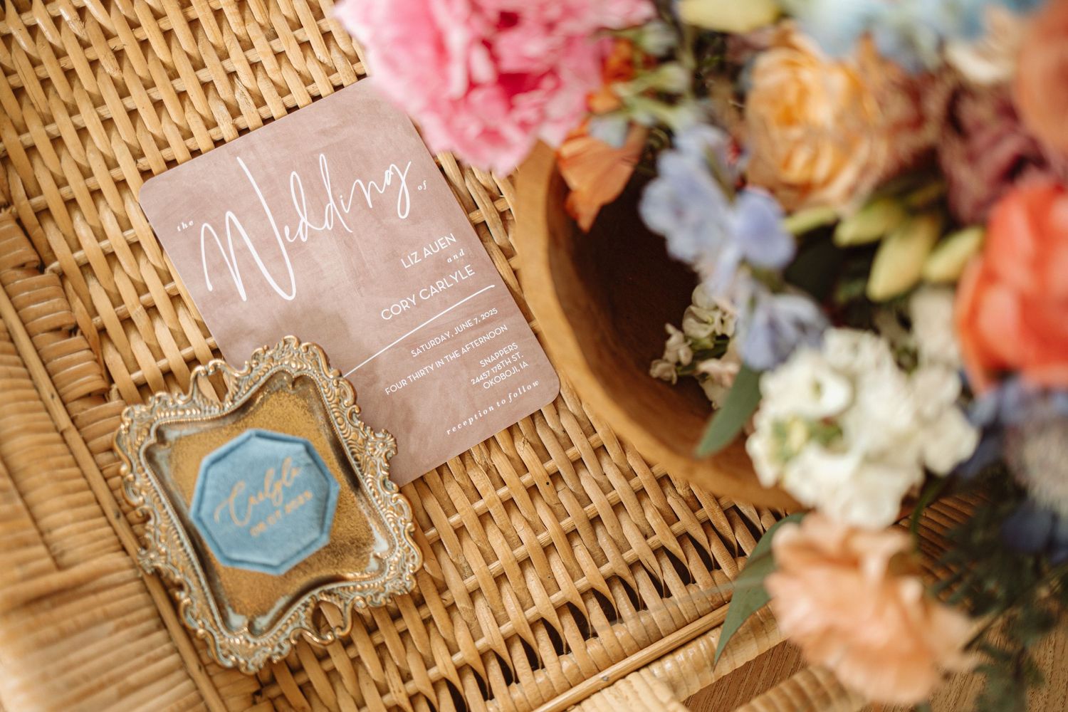 Wedding invitation and blue decorative monogram displayed on woven surface with colorful spring flowers in the background.