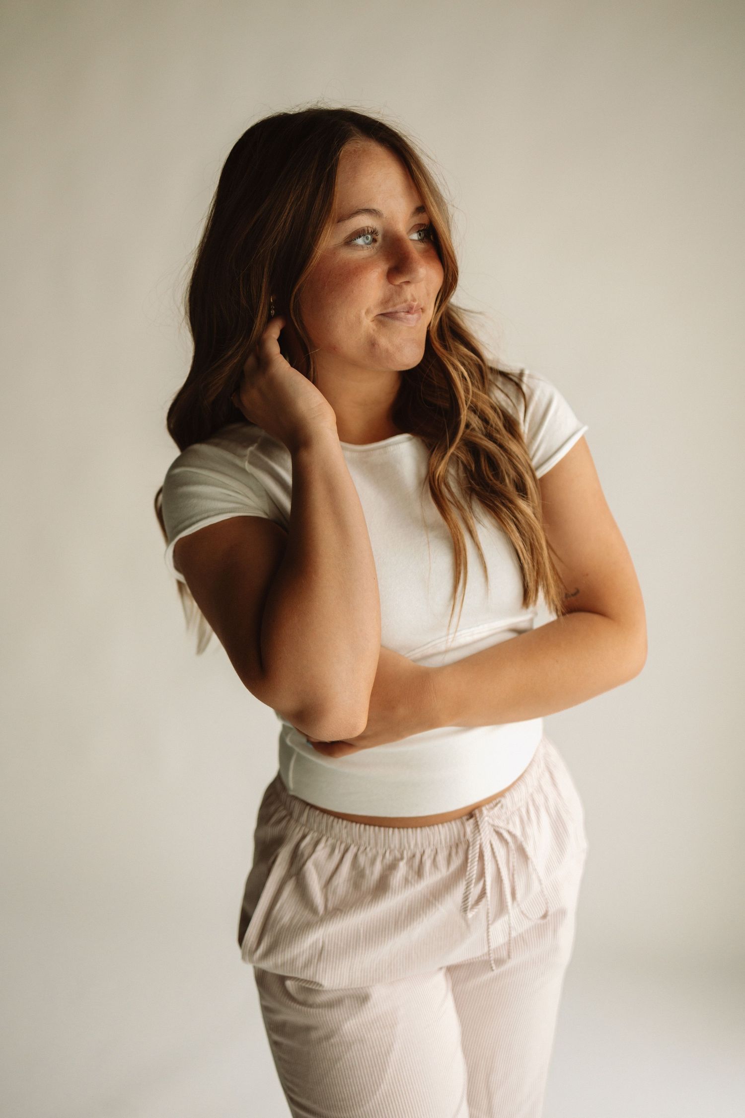 Woman in casual white top and beige pants poses against neutral background with hand in hair and soft smile.