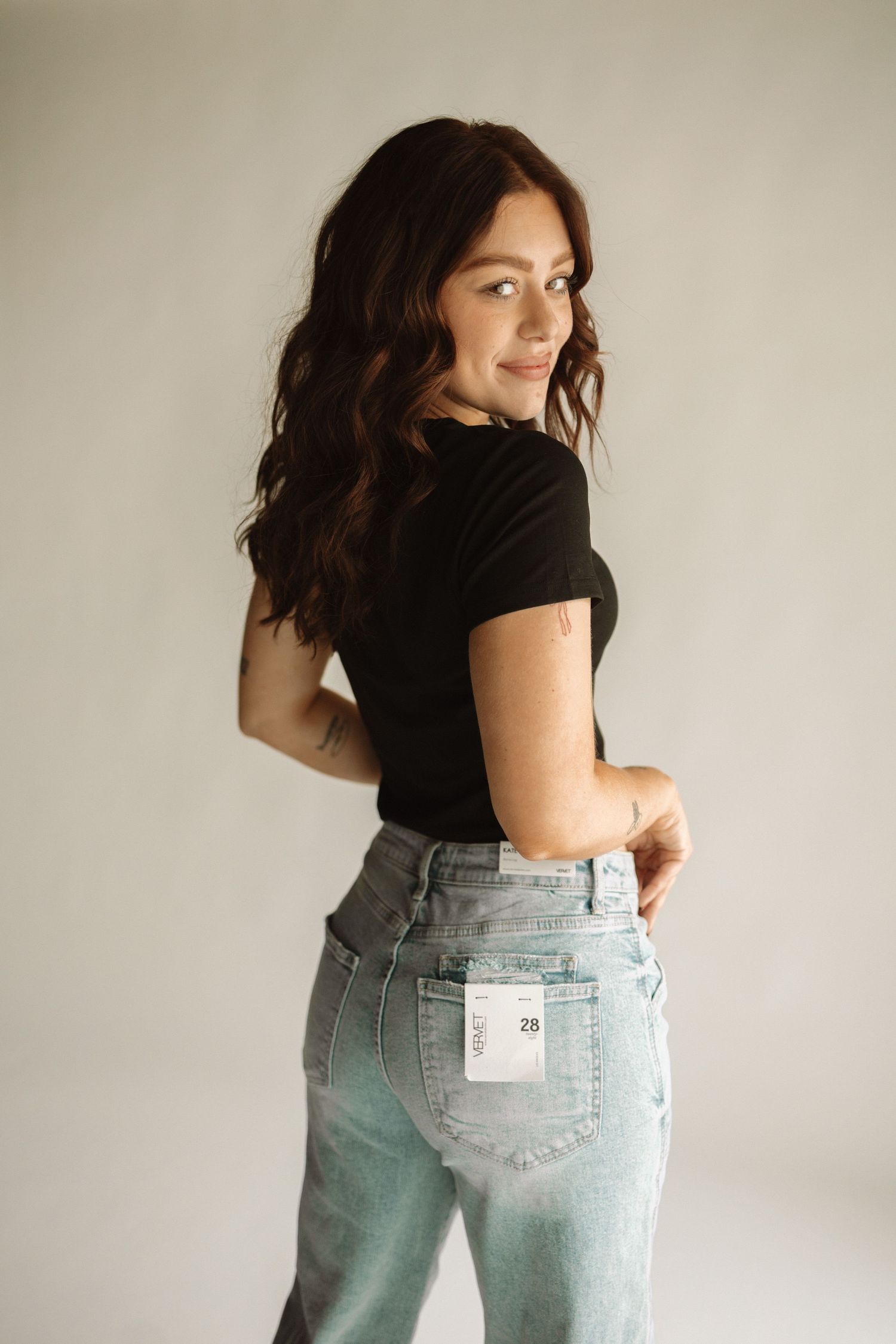A person poses in casual attire wearing a black t-shirt and light wash denim jeans against a neutral background.