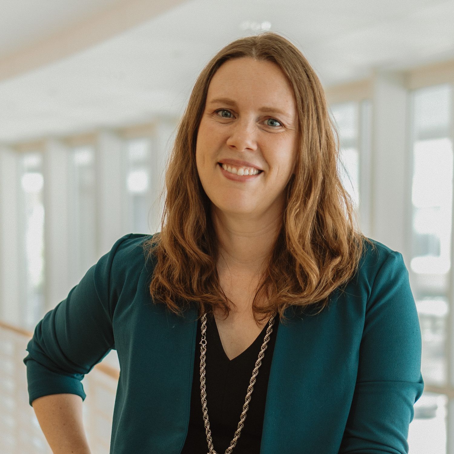 Professional headshot of someone wearing a teal blazer and long necklace in a bright office setting.