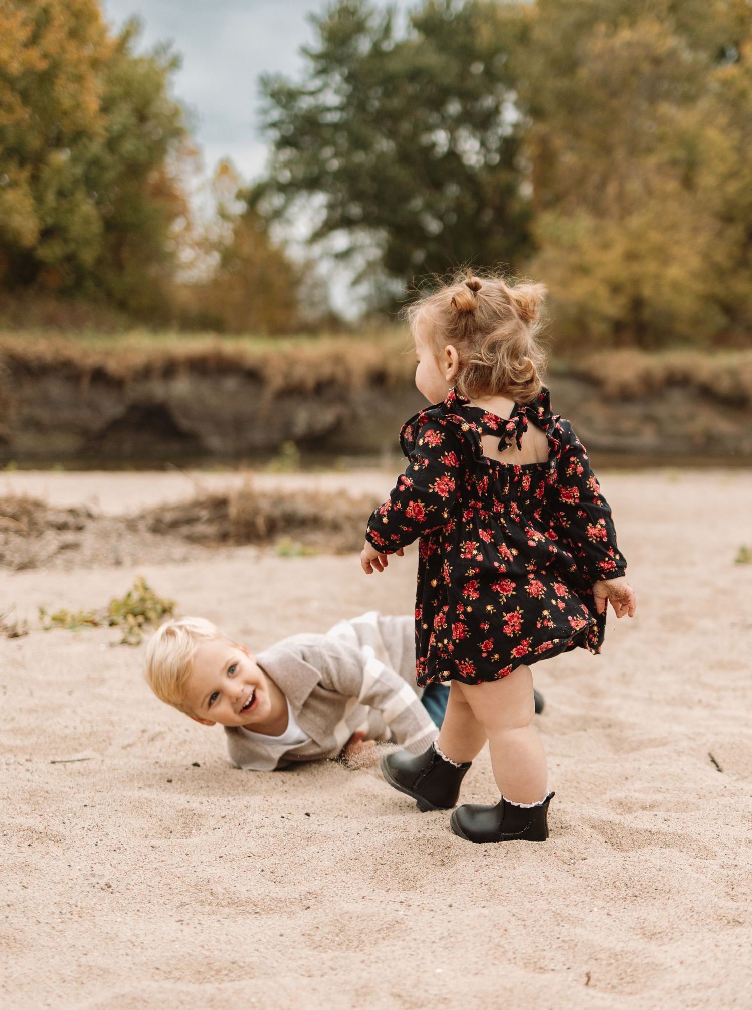 Two young children playing and laughing together on sandy ground with trees in the background.