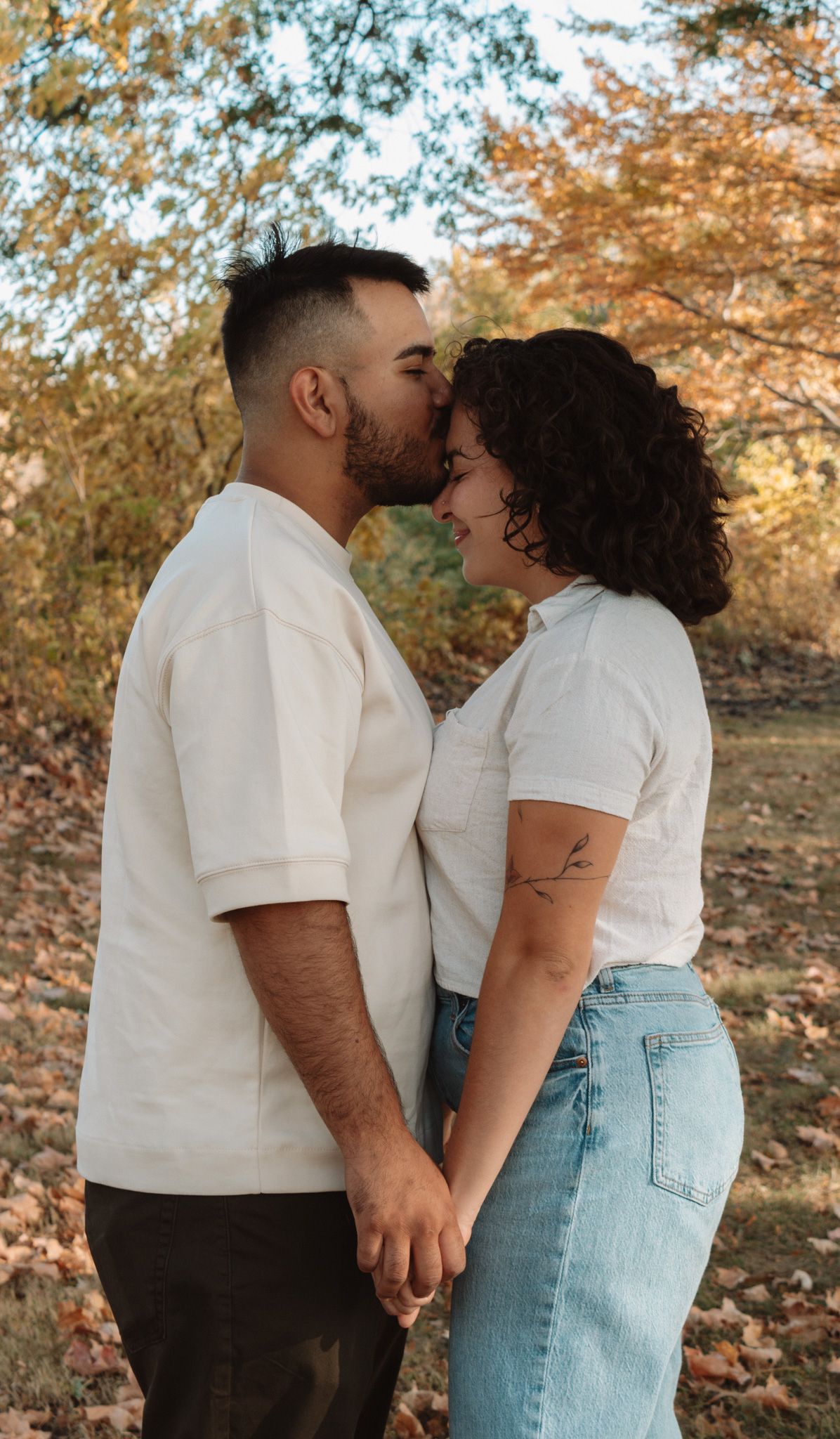 A couple shares an intimate moment in a fall setting with golden leaves and sunlight filtering through the trees.