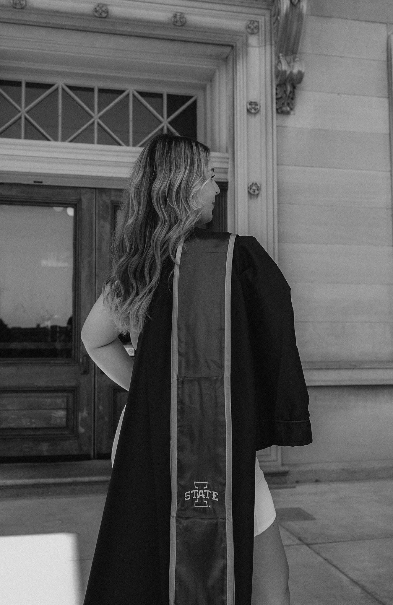 Graduate in commencement gown stands in front of ornate building entrance.