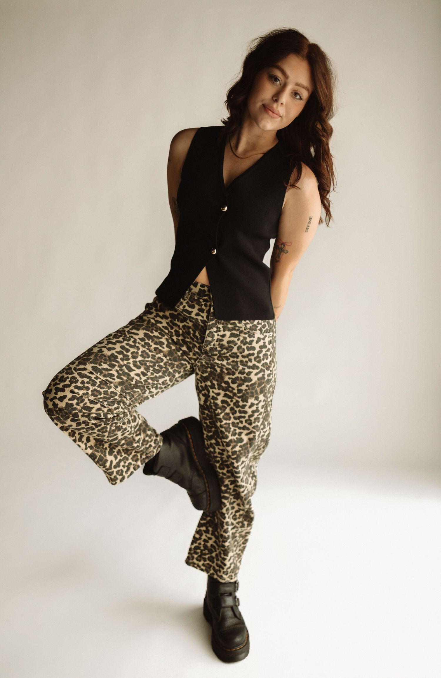 Fashion model poses in black vest top and leopard print pants with black boots against white studio background.
