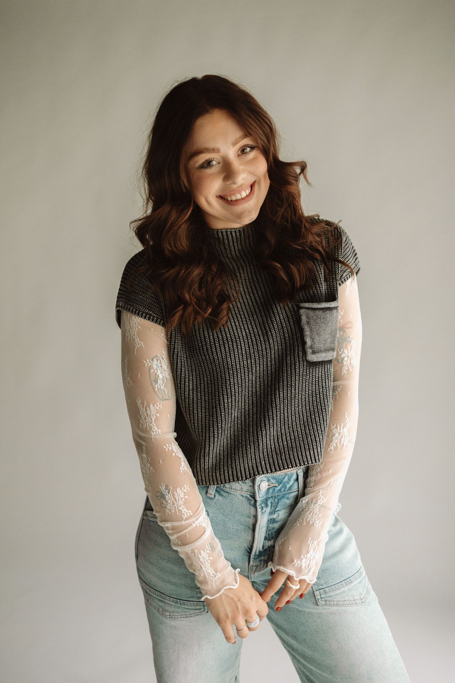 Model wearing gray ribbed sweater vest with white lace sleeves and light blue denim jeans against neutral background.