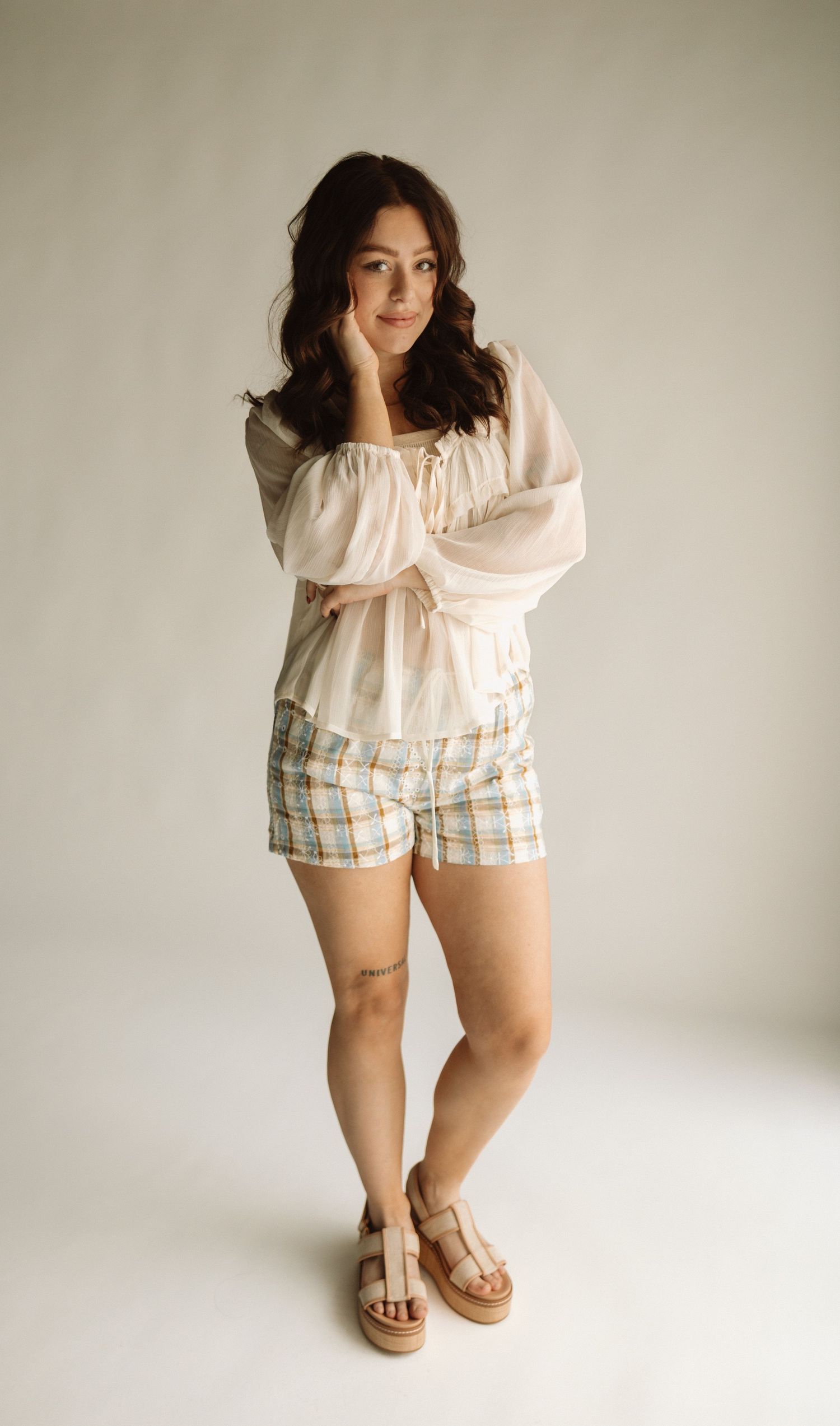 A fashion model poses in a light pink blouse, plaid shorts, and platform sandals against a neutral background.