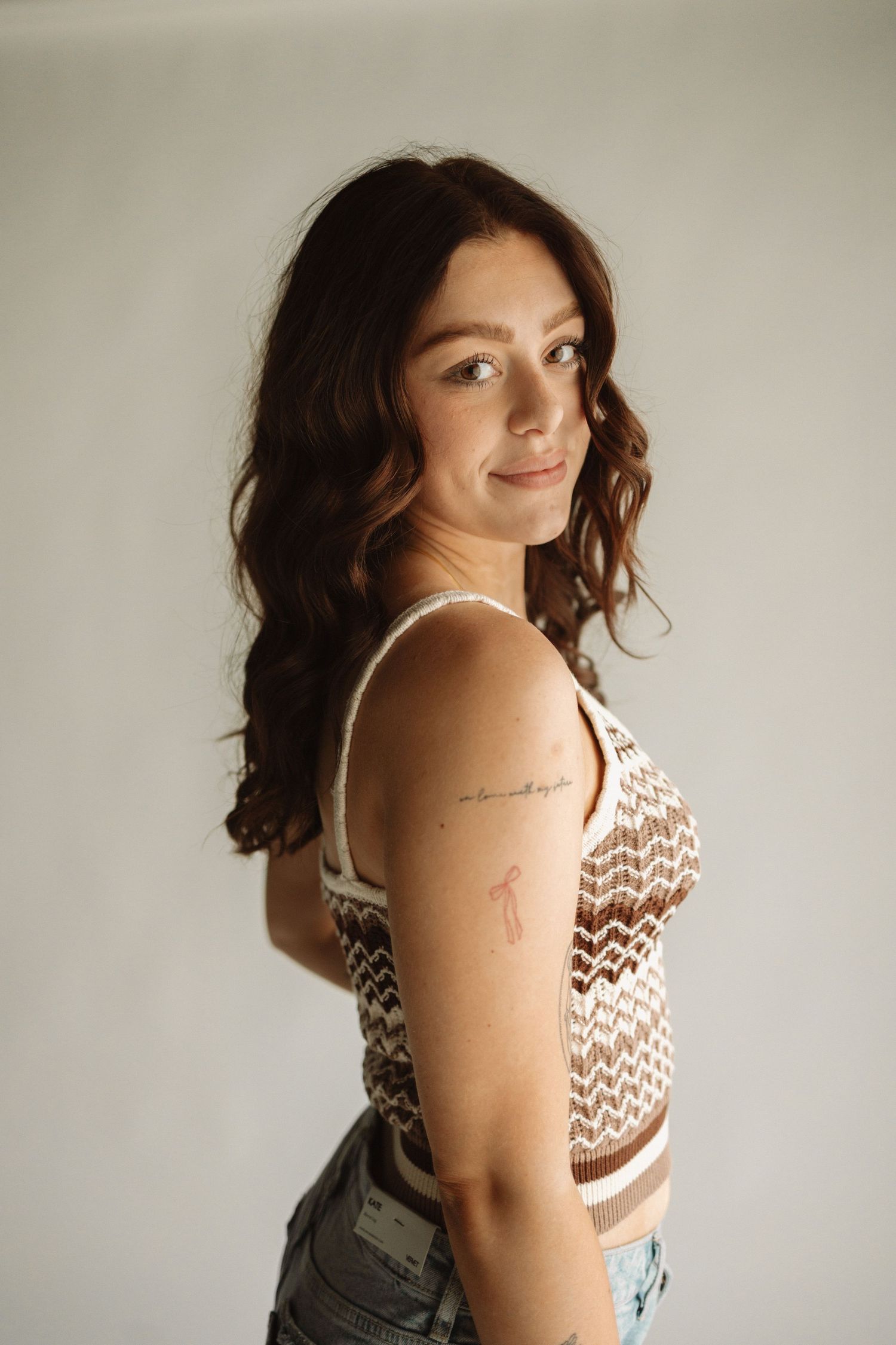 Side profile portrait against white wall wearing chevron patterned tank top and denim, with delicate arm tattoo visible.
